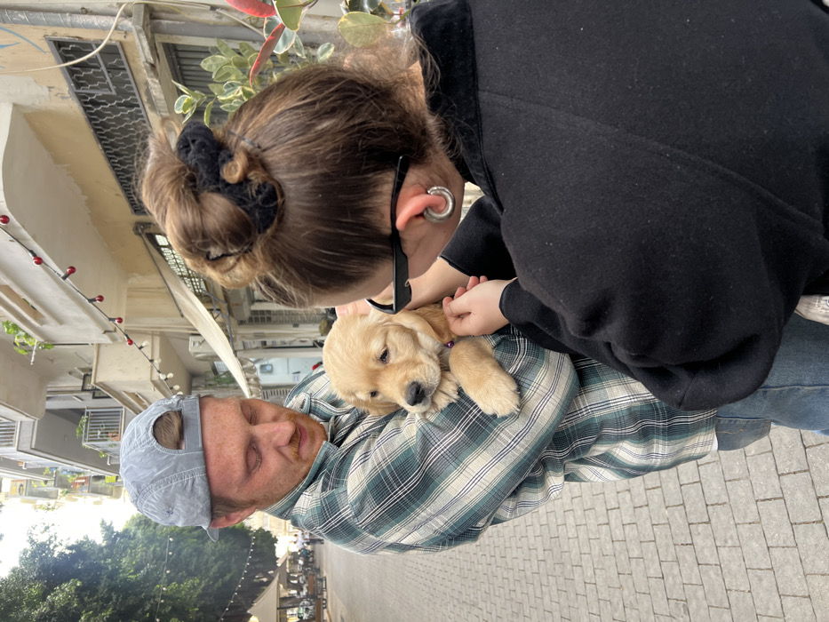 An outdoor urban scene depicts two individuals and a puppy on a pedestrian street in Tel-Aviv, Israel, resembling central districts such as Nachalat Binyamin or Sheinkin Street.
A light-skinned male, with reddish hair and stubble, wears a reversed light grey baseball cap, a green and white plaid long-sleeved shirt, and blue jeans. He cradles a light golden-colored golden retriever puppy in his left arm, looking down at it. The puppy wears a thin purple collar and appears calm.
Standing to the male's right, a female is seen primarily from behind. She has brown hair styled in a messy bun secured with a black scrunchie. Black-framed sunglasses rest on her head/ears. She wears large silver hoop earrings, specifically gauges, in both visible earlobes and a dark, possibly black, coat or jacket. Her hands are positioned near the puppy's head and neck.
The ground is paved with light-colored, interlocking bricks. In the background, multi-story urban buildings with light-colored facades, balconies, and windows are visible. Some ground-floor establishments feature awnings. String lights with small red bulbs are strung overhead. On the left side of the street, distant trees and outdoor cafe seating with white umbrellas are present. A leafy green plant with red-tinged edges is situated on the right side. The scene is brightly lit by natural daylight.