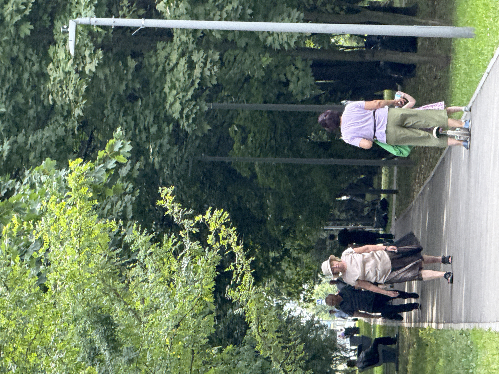 The image depicts a paved pathway in a park, bordered by lush green trees and grass. The pathway is illuminated by streetlights on tall poles. Several individuals are visible on the pathway. In the foreground, a woman wearing a light-colored hat, a beige top, and a dark skirt walks away from the viewer. Behind her, a man in a striped t-shirt and dark pants carries a black bag. Further back, a person is seated on a park bench on the left side of the path, and another person in dark clothing walks away from the viewer on the right. In the far distance, several figures are walking. The lighting suggests it is daytime, with sunlight filtering through the trees. The overall atmosphere is that of a tranquil park setting.