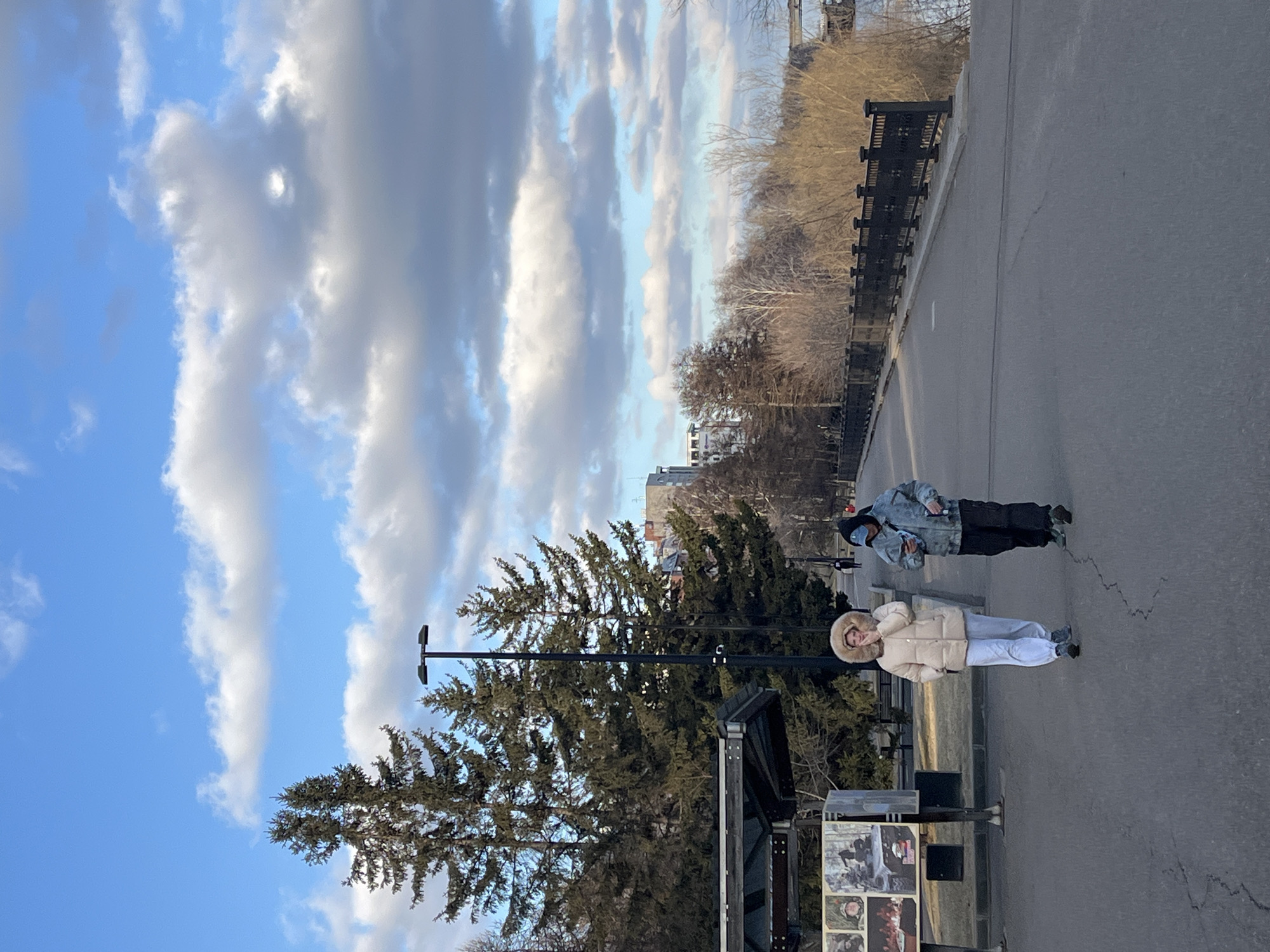 The image captures an outdoor scene in what appears to be an urban park or embankment in Krasnoyarsk, Russia. Two individuals are standing on a paved path or road. The person closer to the foreground is wearing a light-colored (possibly cream or beige) puffer jacket with a prominent fur-lined hood, light-colored pants, and dark shoes. Further back, another person is dressed in a darker, potentially camouflage-patterned jacket with a hood up, dark pants, and light-colored footwear.
A large evergreen conifer tree dominates the left side of the frame, with its branches extending towards the top. A black street lamp with two fixtures stands between the people and the tree. To the right of the person in the light jacket, there is an information stand or kiosk displaying multiple framed images, which appear to be black-and-white historical photographs. Any accompanying text below these images is too small to be legible.
The setting includes a paved ground surface, which shows visible cracks. In the background, beyond the paved area and a fence or railing, are numerous bare deciduous trees, suggesting a late autumn, winter, or early spring season. Distant city buildings are faintly visible through the trees. The sky above is a vibrant blue, partially covered by large, dramatic white and grey clouds. The overall light suggests daytime, possibly late afternoon given the quality of light on the clouds. The individuals appear to be casually standing or observing their surroundings.