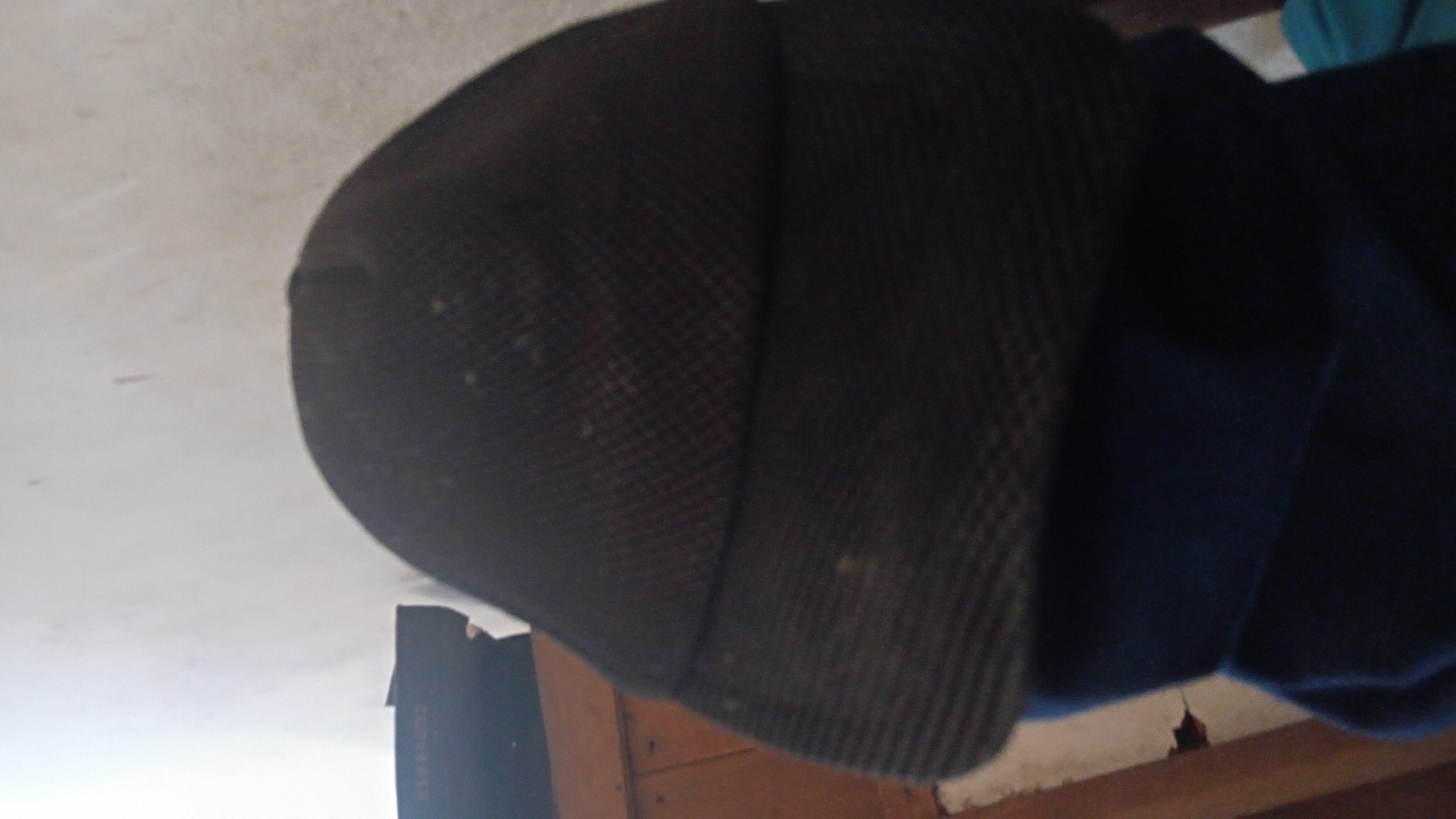 A close-up, low-angle shot shows a dark-colored baseball cap resting on top of a dark blue garment, likely a shirt or jacket. The cap has a textured surface, appearing to be made of a mesh-like material, and a brim. The background consists of a plain, lightly textured wall and a wooden surface on the left, possibly a piece of furniture. The lighting is dim, with a slight glare on the wall. The image is blurry and lacks sharp detail. No specific location within Prabumulih, Indonesia can be determined from the visual information provided.