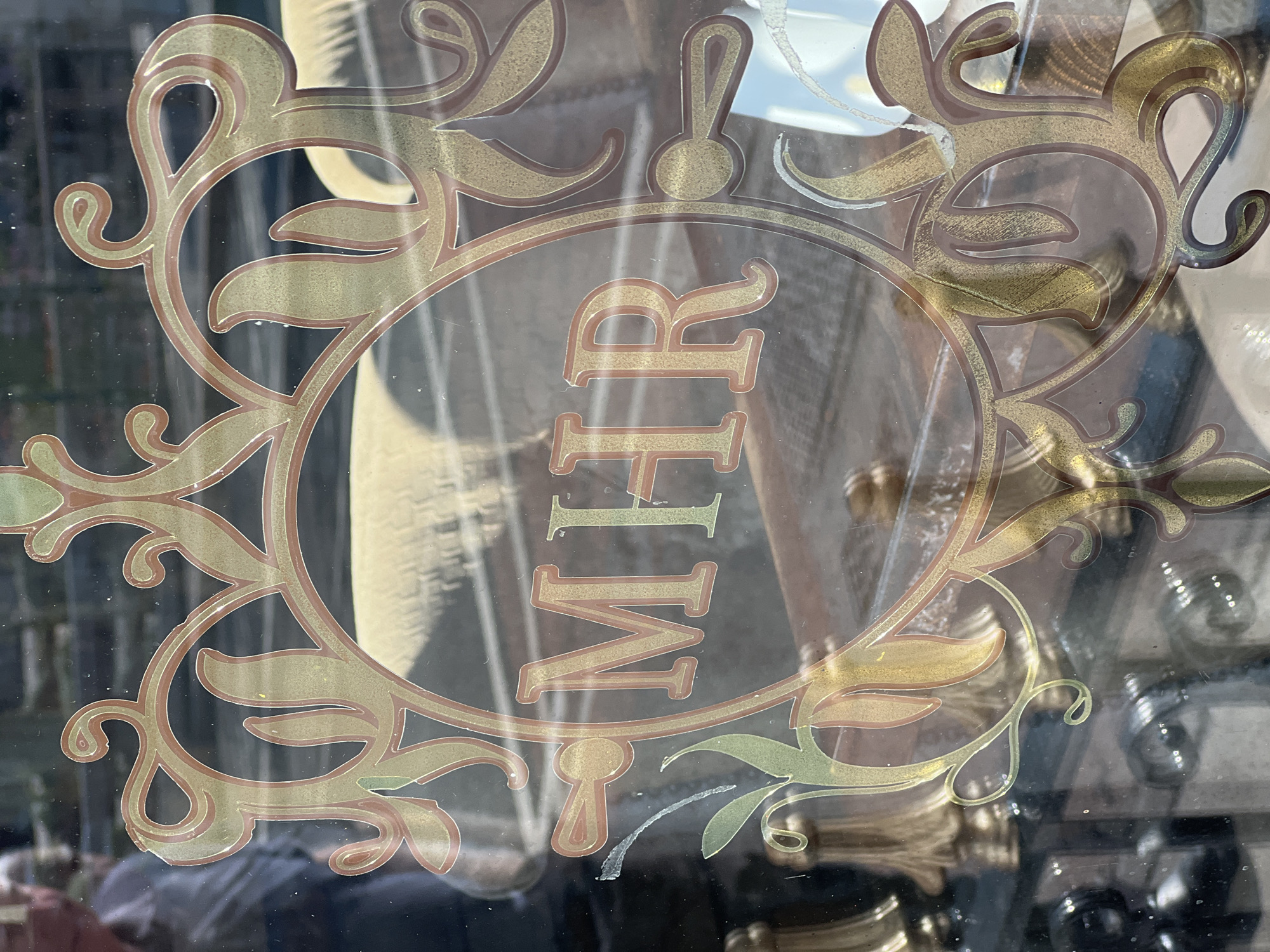 The image is a close-up shot of a glass window with ornate gold and red decorative elements. In the center, the letters "MHR" are displayed in large, stylized gold lettering within a circular motif. The glass reflects a blurry background, which appears to be an indoor setting with furniture. The decorative elements consist of swirling lines, leaf-like shapes, and what seem to be keyhole or lock-like designs, all rendered in a metallic gold with a reddish-brown outline. The window is likely part of a shop or establishment in Donegal, Ireland, given the presence of the letters "MHR" which may represent the initials of the business.