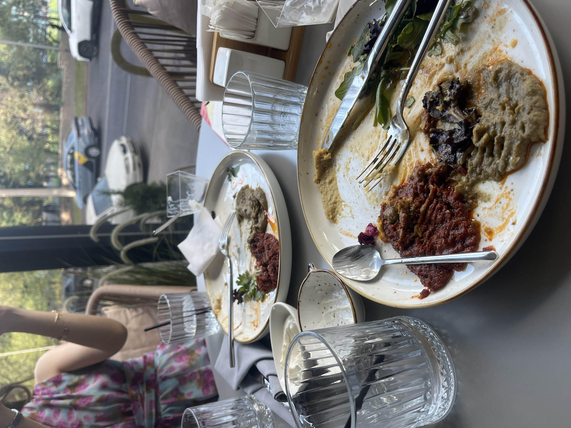 A close-up, eye-level shot captures a table setting at what appears to be a restaurant in Almaty, Kazakhstan. The scene is dominated by two partially eaten plates of food, with remnants of dips, vegetables, and sauces scattered across their surfaces. Utensils, including a fork and a spoon, rest on the plates. Several clear glasses, some containing water and ice, are also present on the table. A woman in a floral dress is partially visible in the background, her arm extended. Outside the window, traffic on a street is visible, including cars and a taxi, suggesting an urban environment. The overall impression is that of a meal that has recently concluded, with the remnants left behind.