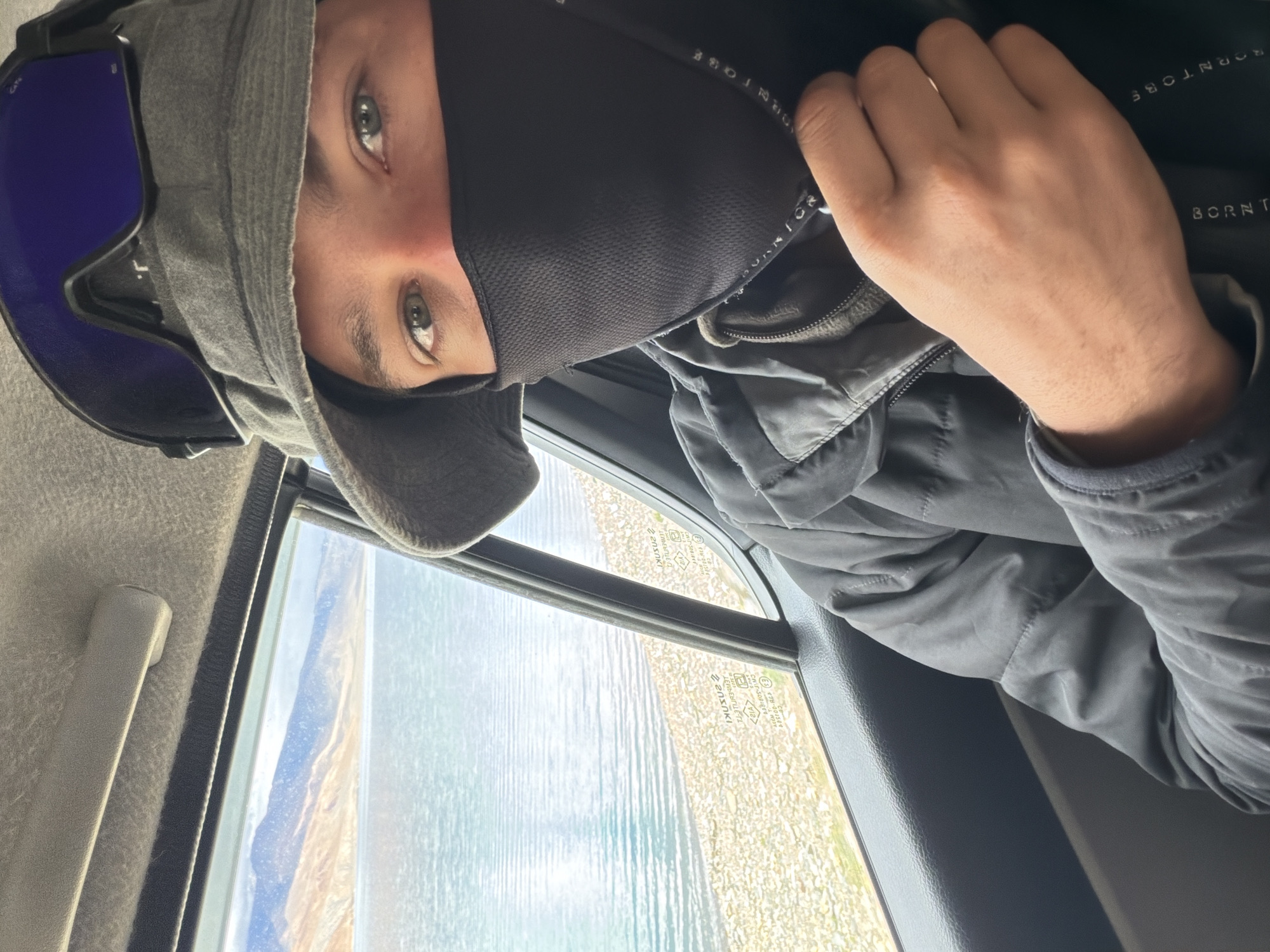 A selfie taken inside a vehicle shows a person wearing a grey bucket hat and ski goggles, with a black face mask covering the lower half of their face. The person is looking at the camera with their eyes visible. They are wearing a dark jacket. Through the window of the vehicle, a large body of water, possibly a lake, is visible, with hills or mountains in the background. The location is identified as Karzok, India. The interior of the vehicle, including the car door and ceiling, is also visible.