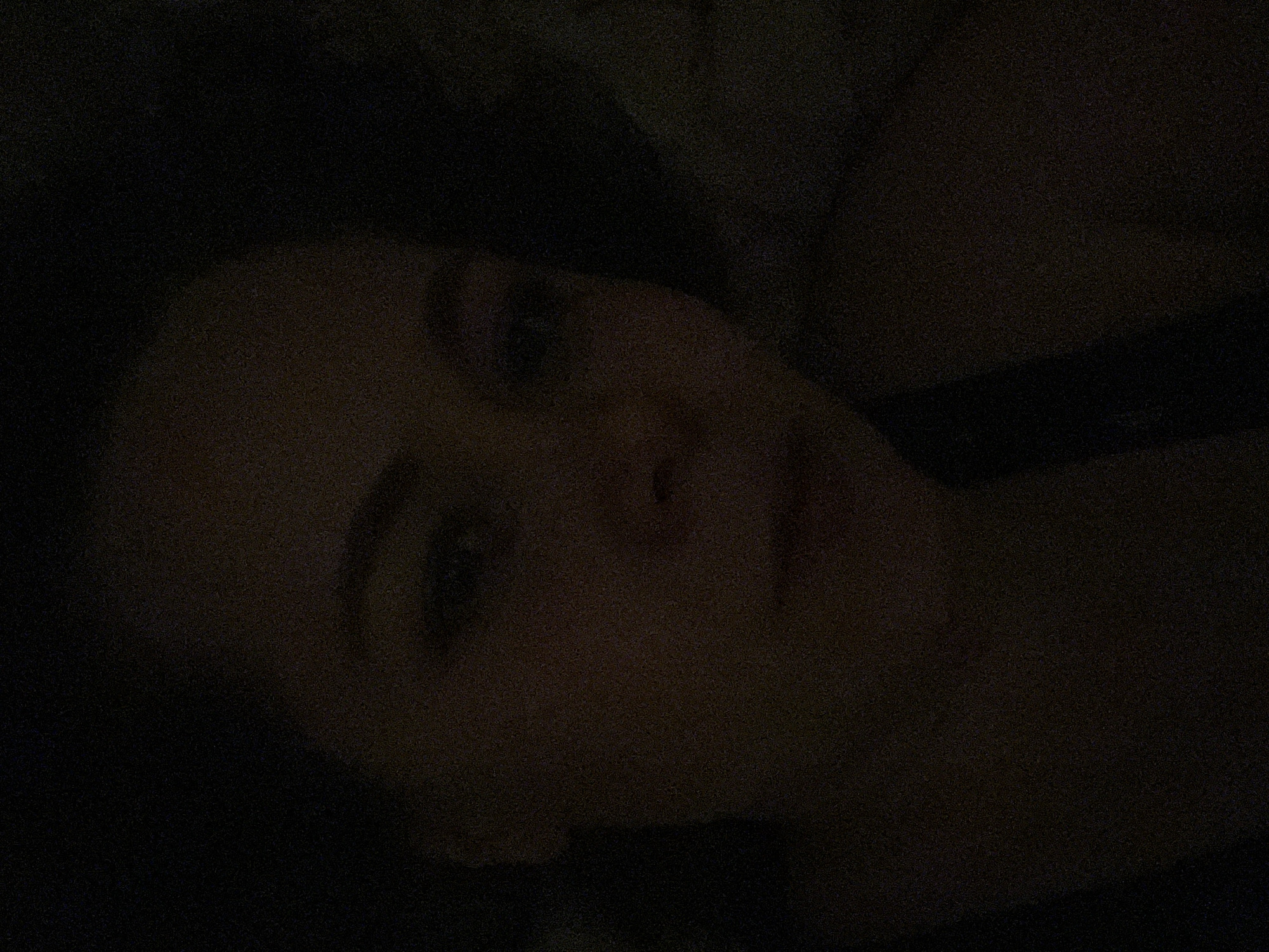 The image shows a close-up, underexposed view of a person's face. The person is looking directly at the camera with their head tilted slightly to the right. The person's right shoulder and a portion of a black strap are also visible. The location is not discernible from the image.