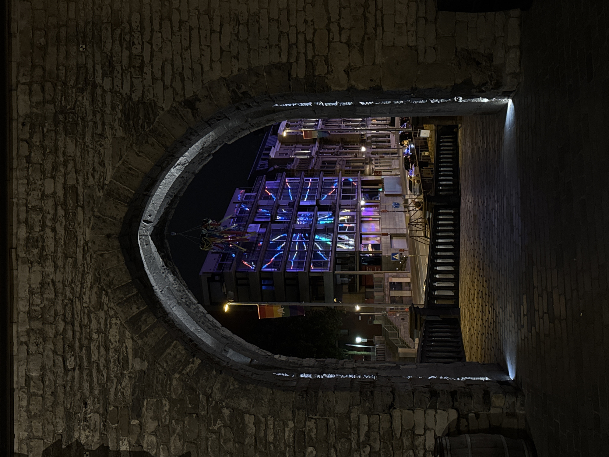 This is a nighttime, eye-level, medium shot photograph taken through an ancient stone archway in Antwerp, Belgium. The archway, made of large, rough-hewn stones, frames a view of a modern building across a street. The building features a facade with numerous windows illuminated by colorful, abstract light patterns in blues and purples. A rainbow-colored flag hangs from a lamppost in front of the building. The foreground consists of a brick-paved walkway leading through the archway, with small spotlights embedded in the stonework illuminating the path. A wooden barrel is visible to the left of the archway. Cars and a few pedestrians are present on the street beyond the archway. The overall atmosphere is urban and festive.