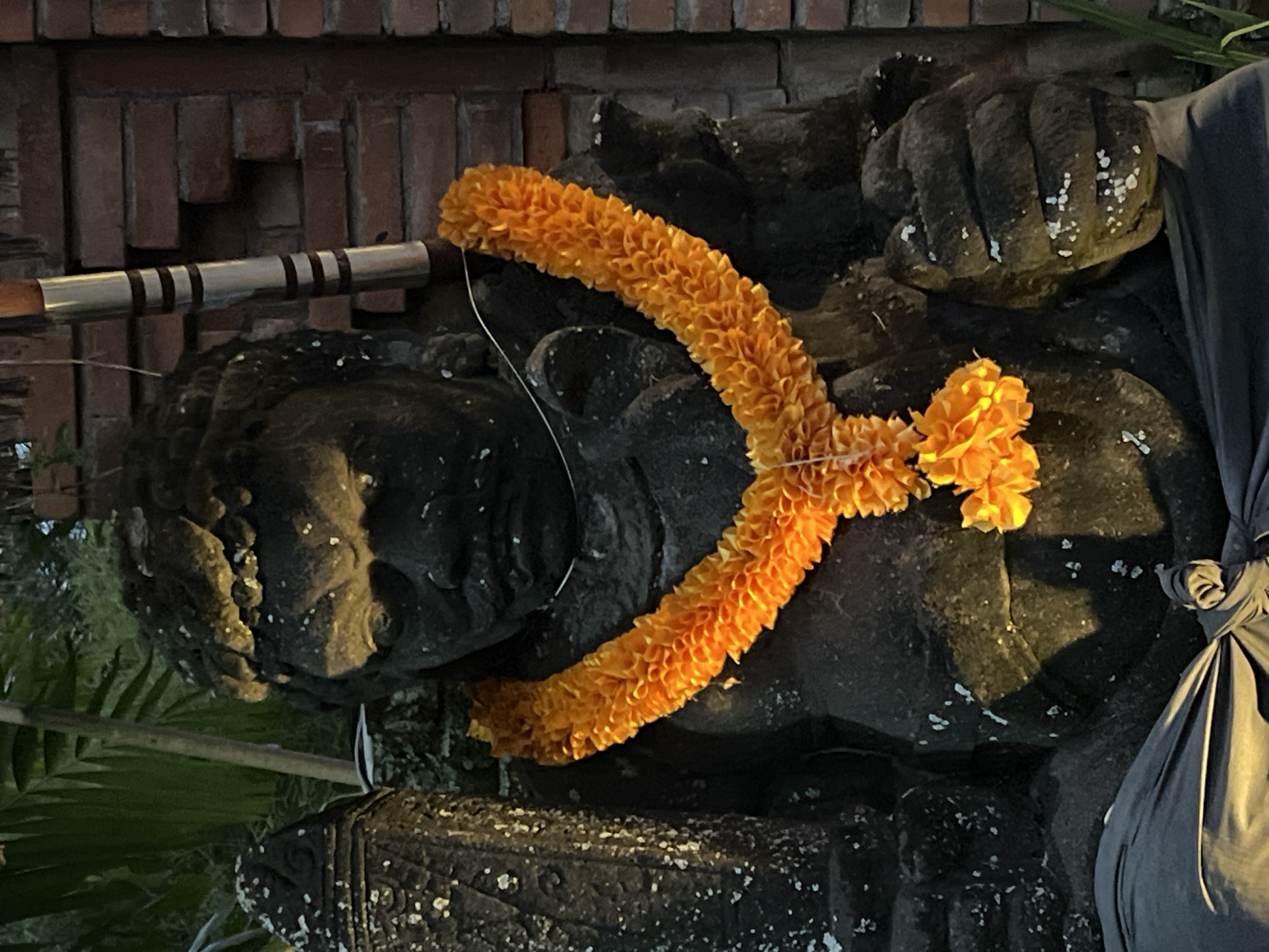 A horizontal, medium-shot photograph shows a stone statue of a figure adorned with an orange flower garland, likely located in the Ubud District, Indonesia. The statue, rendered in dark grey stone, is positioned against a background of brickwork. An orange flower garland is draped over the head and shoulder of the figure. A metal pole with dark bands is visible behind the statue. The statue is partially draped with fabric on the right. Greenery is visible at the bottom and top right corners of the image.
