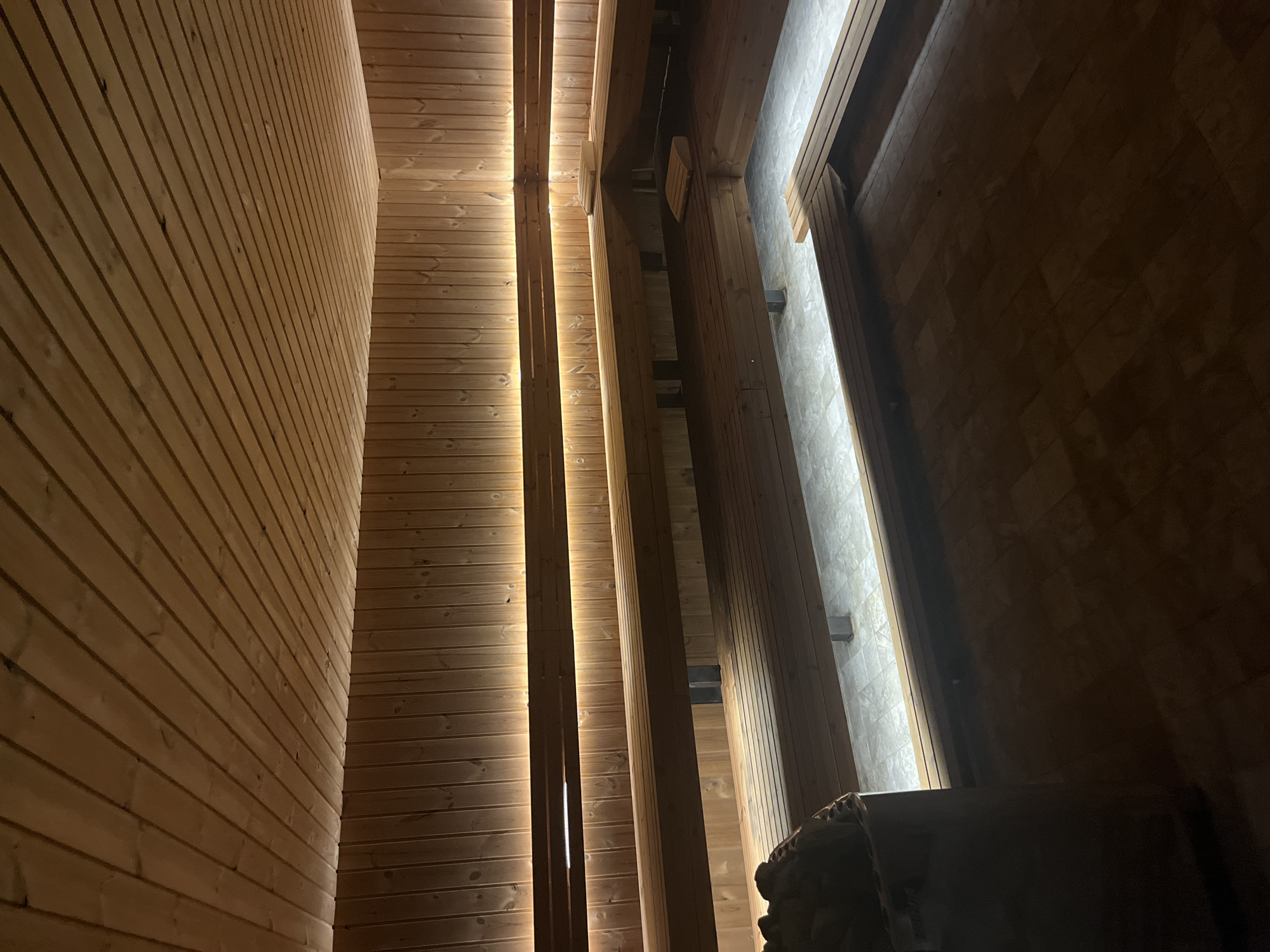 The image depicts the interior of a sauna, likely located in Sofia, Bulgaria, given the user's location. The sauna is constructed with light-colored wood paneling. The left side of the frame features a curved wooden wall with horizontal planks. Vertical wooden beams are placed at intervals, with recessed lighting between the panels and beams, creating a glowing effect. On the right side, there is a corner with wooden beams and a lighter-colored, possibly tiled, wall. In the lower right corner, a dark, indistinct object that could be a sauna heater or stones is present. The scene is dimly lit, with light sources being primarily the recessed lighting and indirect light from the tiled wall. No people are visible.