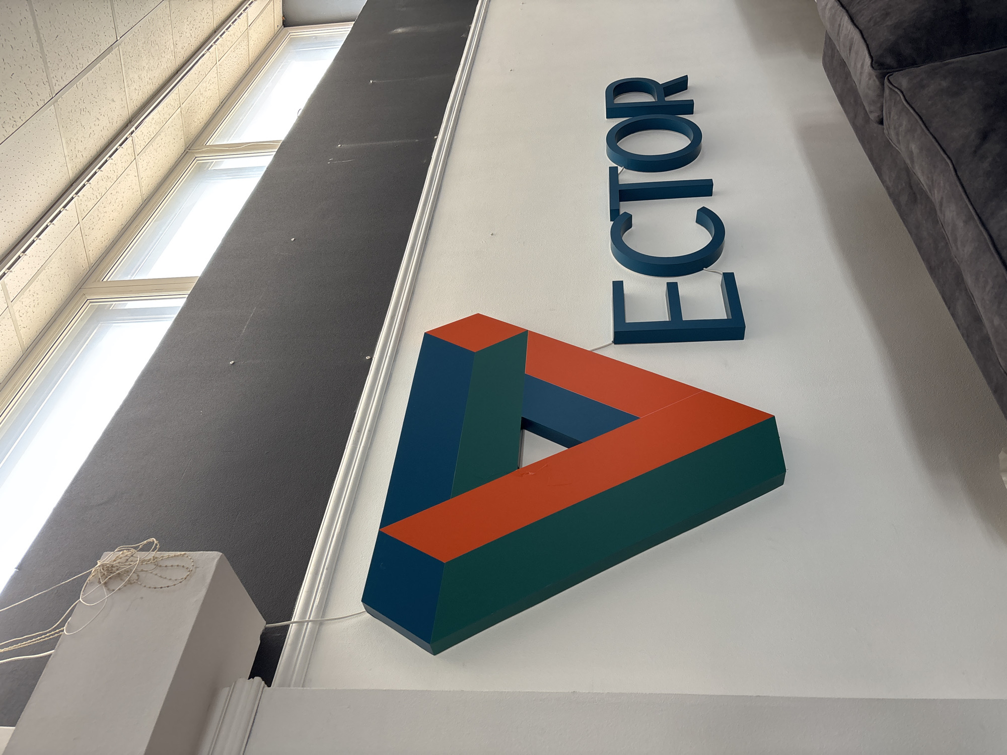 In Zaporizhzhia, Ukraine, a 3D logo is mounted on a white wall. The logo, shaped like an impossible triangle, features teal and orange segments. To the right of the logo, the word "ECTOR" is spelled out in teal block letters. Above the logo and the wall is a dark gray textured panel, and above that, a row of fluorescent lights is visible along the ceiling. A light-colored wire hangs from the top left corner of the frame. In the foreground, the tops of a dark gray upholstered sofa are visible.