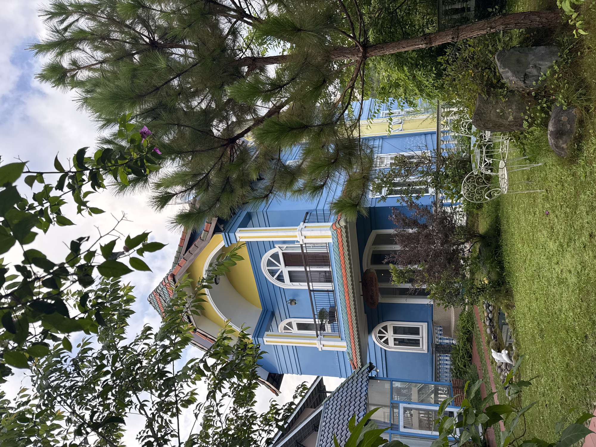 This is a full, eye-level shot of a vibrant, blue two-story house in Lâm Viên - Đà Lạt, Vietnam. The house features a yellow trim and a multi-colored tiled roof with shades of red, orange, green, and gray. A balcony with a black metal railing extends from the second floor. The front entrance of the house has a sign that reads "Rainbow Da Lat." White decorative chairs and a table are set up on the green lawn in front of the house. A small pond with a white bird statue is also visible in the foreground. The yard is lush with green grass, various trees, and bushes. The sky above is partly cloudy.