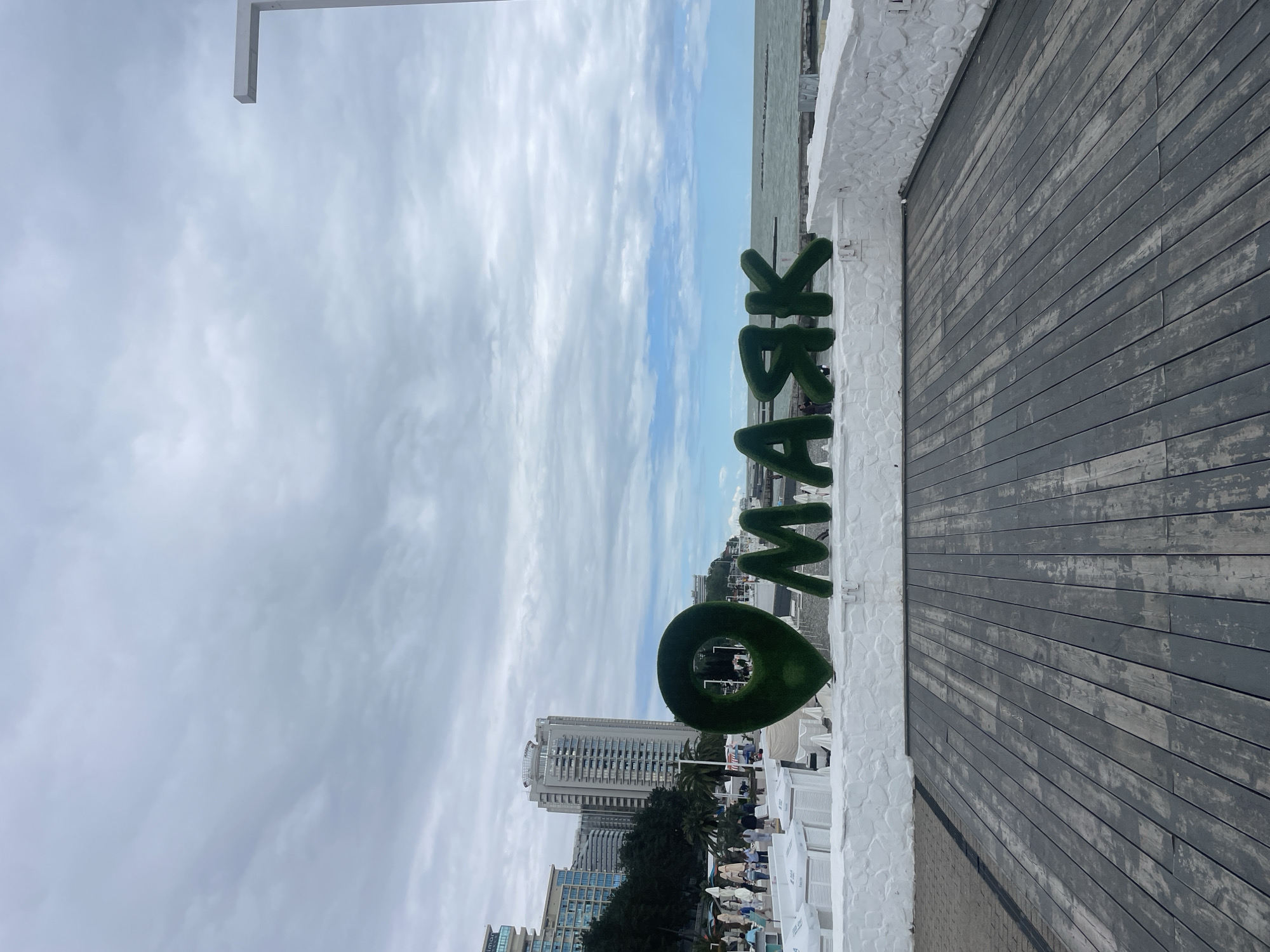 The image shows a view from a wooden boardwalk with a large, green, topiary-style sign spelling "O'WASK" in the foreground. The sign is positioned near a white, textured wall. In the background, there are buildings, trees, and a glimpse of the sea under a cloudy sky. People are seen in the background between the sign and buildings. This appears to be a location in Sochi, Russia.