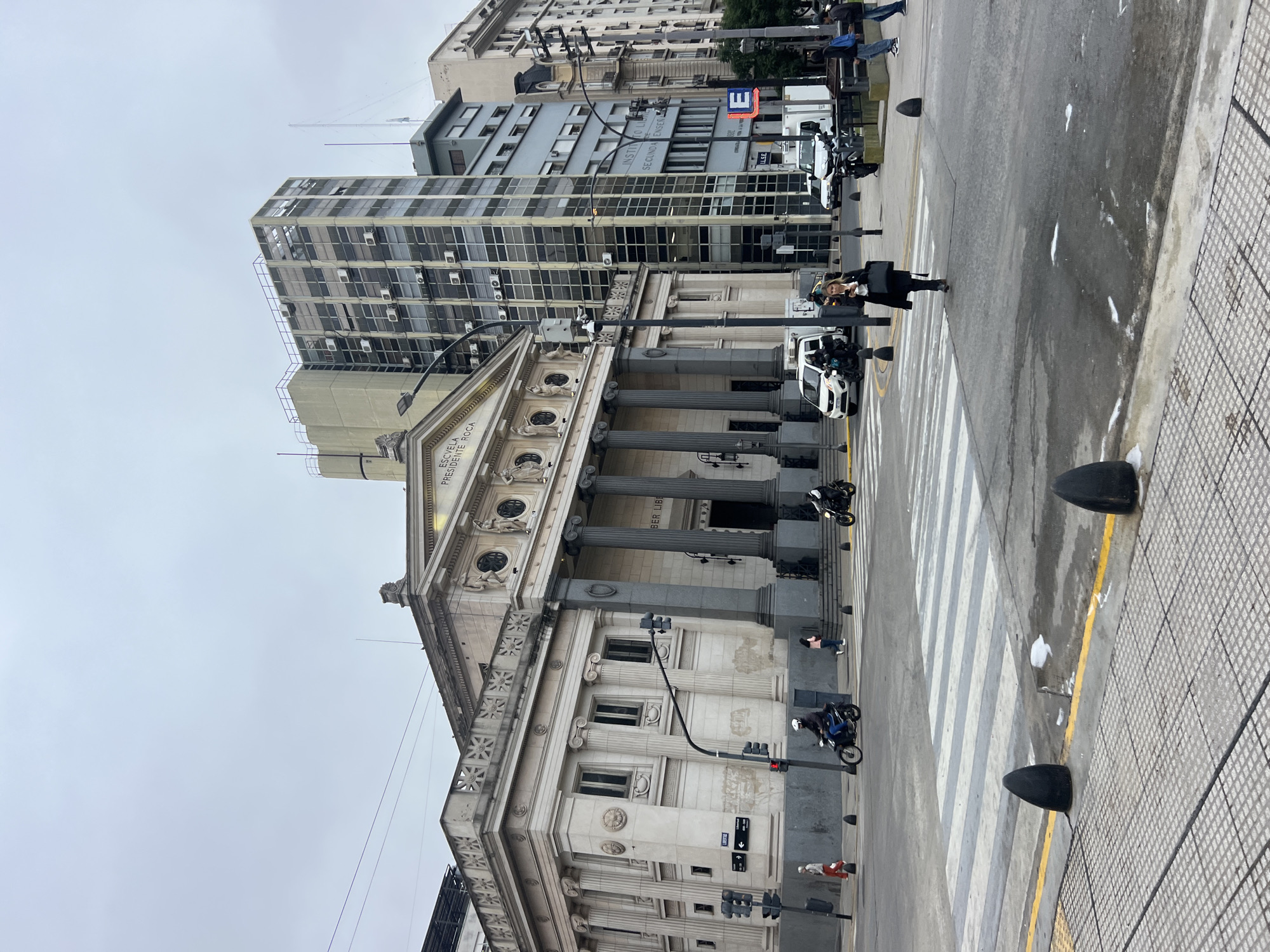 The image depicts the Escuela Presidente Roca in Buenos Aires, Argentina. The building is a classical-style structure with columns and ornate details. In front of the building is a zebra crossing on a street with moderate traffic. Pedestrians are present, including a woman crossing the street. The weather appears overcast. Tall buildings are visible in the background.