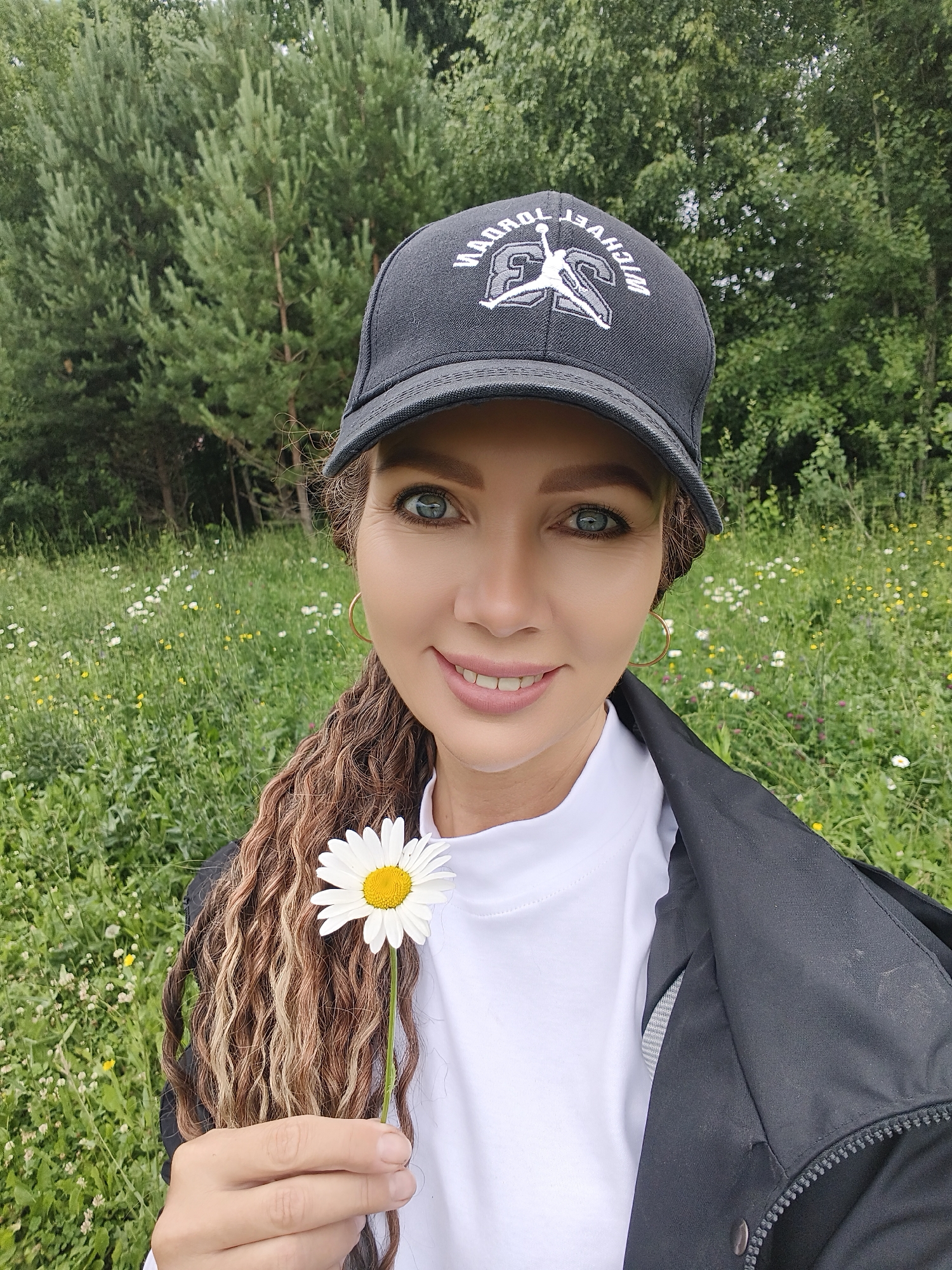 A woman is taking a selfie in a grassy, wooded area. She is wearing a black baseball cap with "MICHAEL JORDAN" and the Jumpman logo embroidered in white and grey. She has long, curly, multi-toned brown hair and is wearing a white turtleneck shirt and a black jacket. She is smiling and holding a single white daisy with a yellow center up to the camera. The background is filled with green trees and a field of wildflowers. The lighting suggests it is daytime, and the weather appears to be pleasant, likely overcast or slightly cloudy, given the soft light. The setting appears to be a natural outdoor environment, possibly a park or a rural area.