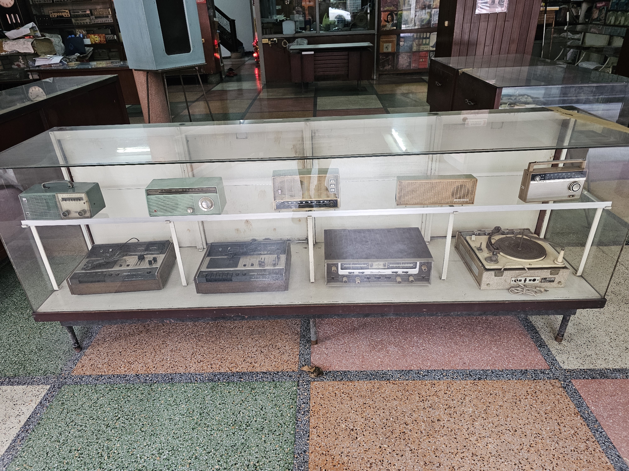 The image displays a collection of vintage electronic devices, including radios, a cassette player, and a turntable, showcased in a glass display case. The devices are arranged on white shelves within the case. Below the display case, the floor is made of terrazzo with a distinctive pattern of colored tiles, including green, orange, and white. In the background, glimpses of what appears to be a shop interior are visible, with shelves stocked with various items, including what look like records. The lighting suggests an indoor setting. The floor pattern, specifically the combination of terrazzo colors and geometric divisions, is characteristic of the flooring found in some older establishments in Bangkok, Thailand. One of the radios has a "SONY" logo.