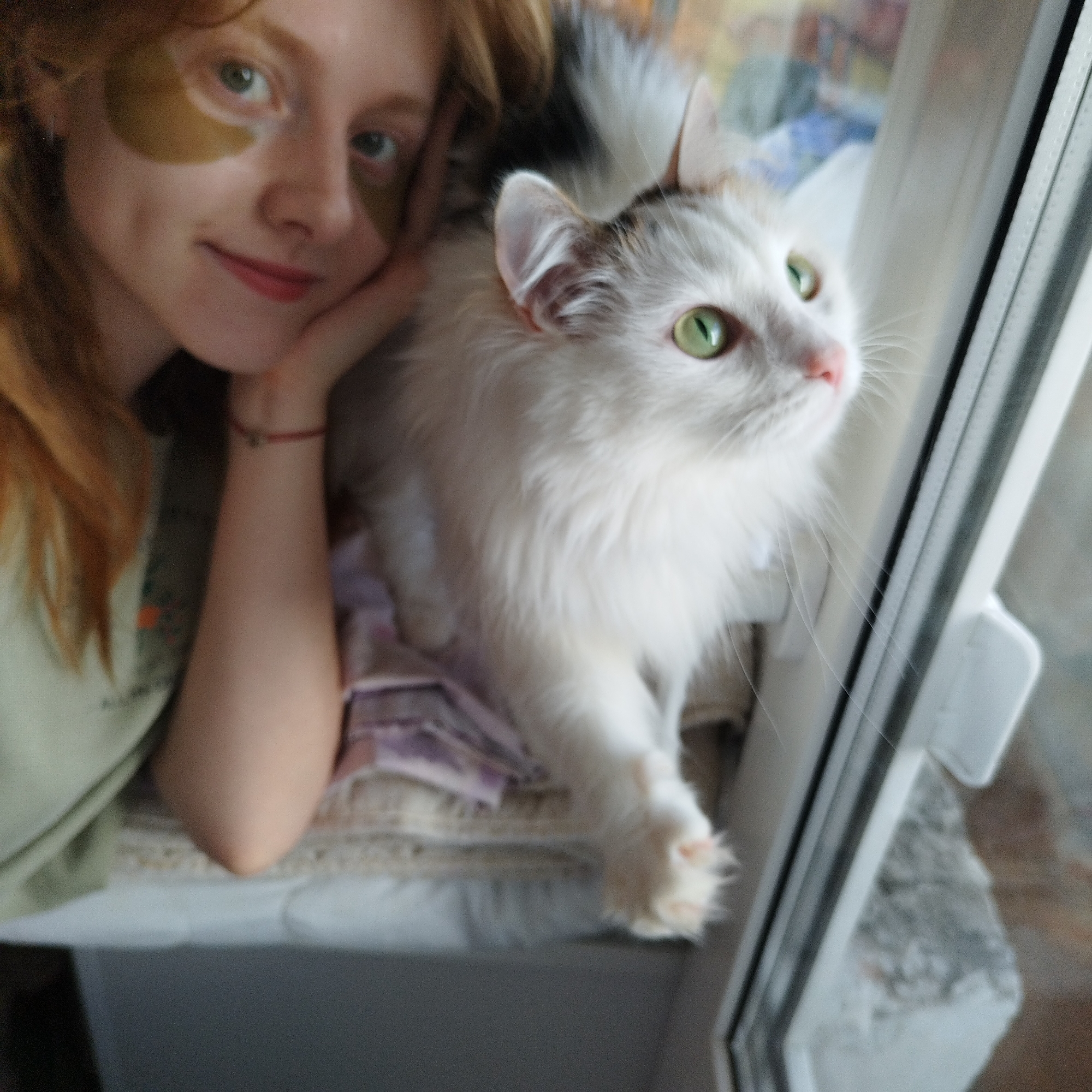 The image features a young woman and a white, fluffy cat positioned indoors, likely by a window. The woman, with reddish-brown hair, is on the left, smiling gently at the camera. She wears green under-eye patches and has a thin red string bracelet on her left wrist, with her chin resting on her hand in a relaxed pose.
Beside her, on the right, is a medium-sized white long-haired cat with prominent green eyes. The cat appears to have some darker, possibly grey or black, markings on its head and tail. It is looking intently out the window to the right, appearing curious or observant of something outside.
Both subjects are situated on what appears to be a windowsill, covered with a patterned, light-colored fabric or cushion. A white window frame is visible on the right side of the image, outlining a blurred outdoor background, which suggests a daytime setting with natural light. The overall mood is calm and suggests an affectionate moment between the woman and her pet. There is no visible text in the image.