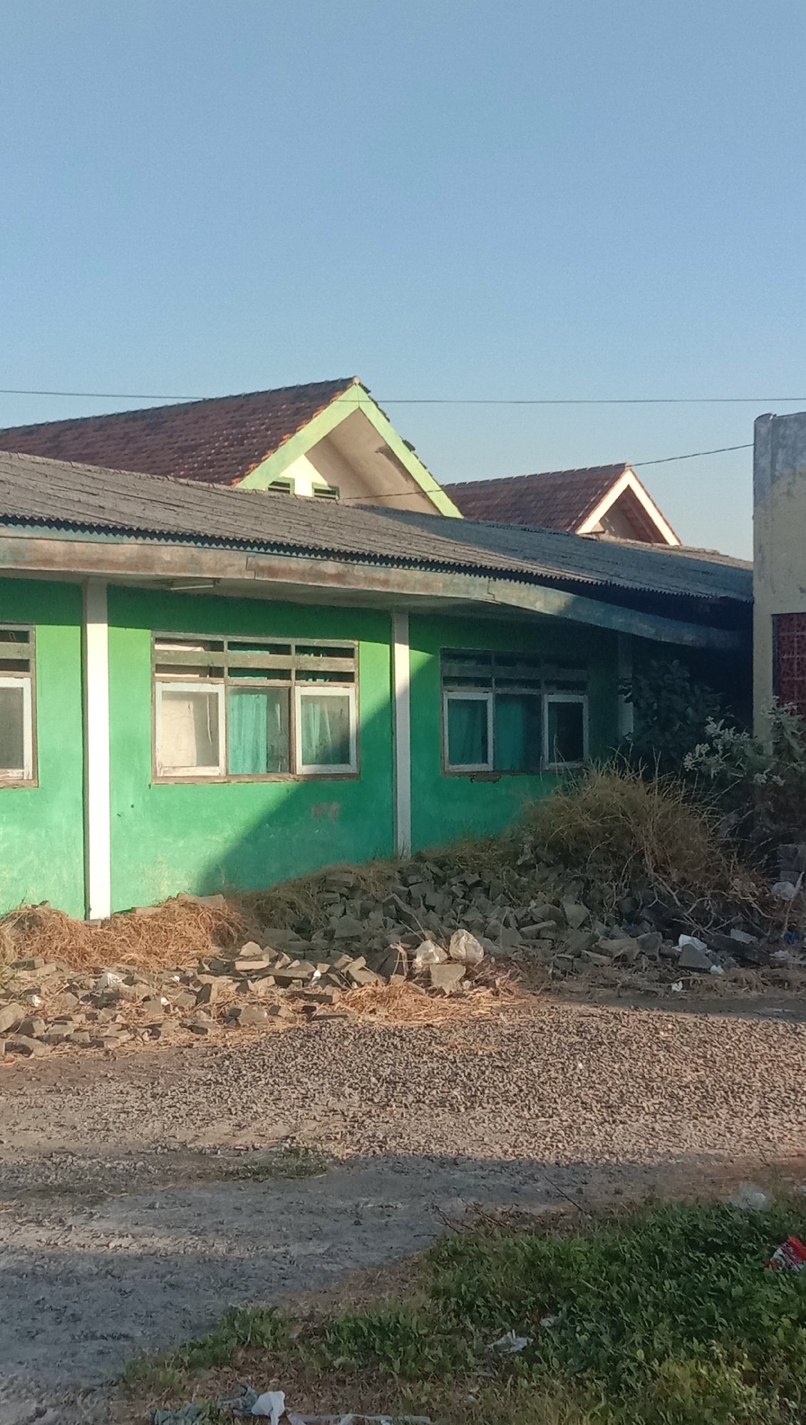 The image depicts a low-angle view of an abandoned or under-construction building with a vibrant green exterior, located in Curah Dukuh, Indonesia. The building features several windows with white frames and teal curtains. The roof is covered with dark gray tiles. In front of the building, there is a pile of rubble and dry grass, suggesting disrepair or ongoing demolition/construction. The ground in the foreground is covered with gravel and some sparse green vegetation. The sky above is a clear, pale blue, indicating it is daytime. The overall impression is one of neglect and a partially completed or decaying structure.
