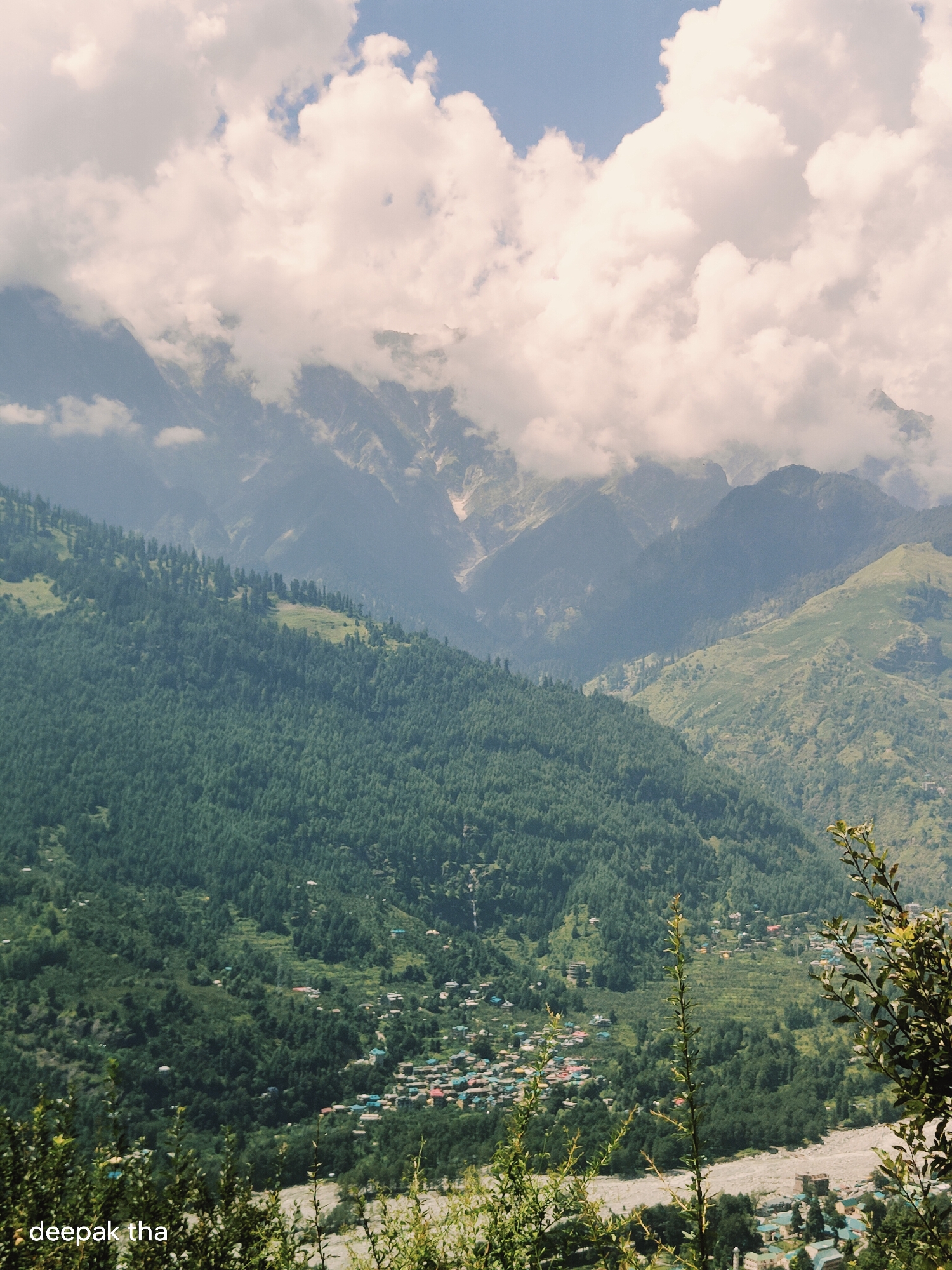 The image shows a panoramic view of a mountainous landscape in Vashisht, India. In the foreground, lush green trees cover the steep slopes, with a small village nestled among them. The village features houses with colorful roofs, clustered along a riverbed. The middle ground is dominated by more dense forests and terraced fields on the hillsides. In the background, majestic mountain ranges rise, partially obscured by dramatic clouds. The sky above is a mix of blue and white, with the clouds casting shadows over the peaks. The overall scene depicts a serene and natural environment with human habitation integrated into the landscape.