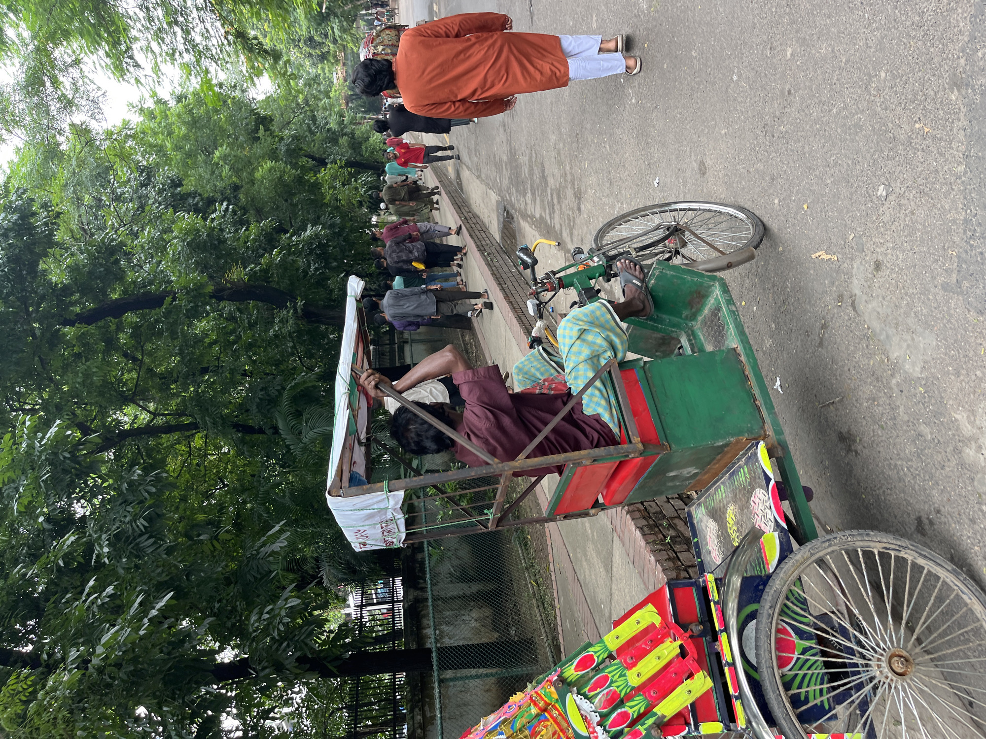 In Dhaka, Bangladesh, a rickshaw driver in a maroon shirt and checkered lungi is seated in his colorful, decorated rickshaw, holding onto the handlebars. He is on a paved road, with a sidewalk to his left. A group of people are walking along the sidewalk, some in the distance. One man in an orange robe and white pants walks away from the viewer on the right side of the road. Lush green trees line the sidewalk, providing shade. The sky is overcast.