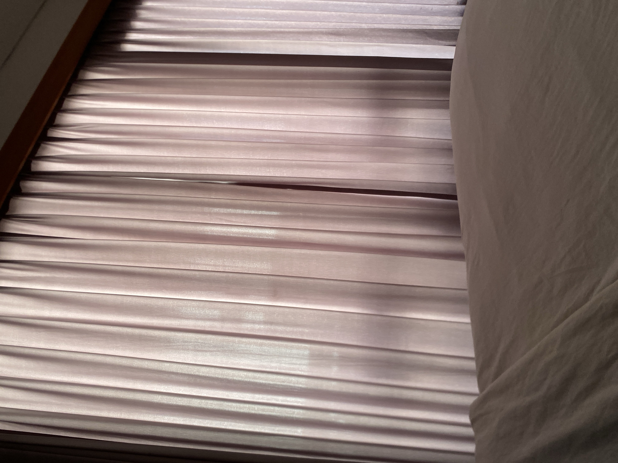 The image shows a set of closed, light purple horizontal blinds filtering sunlight. To the right, there is a blurred, draped white fabric, possibly a bedsheet or curtain. The blinds are installed in a window frame, partially visible on the left side. The scene is indoors and appears to be a bedroom or similar living space. No specific information indicates that the image was taken in North Kuta, Indonesia. Therefore, it's impossible to confirm its location.
