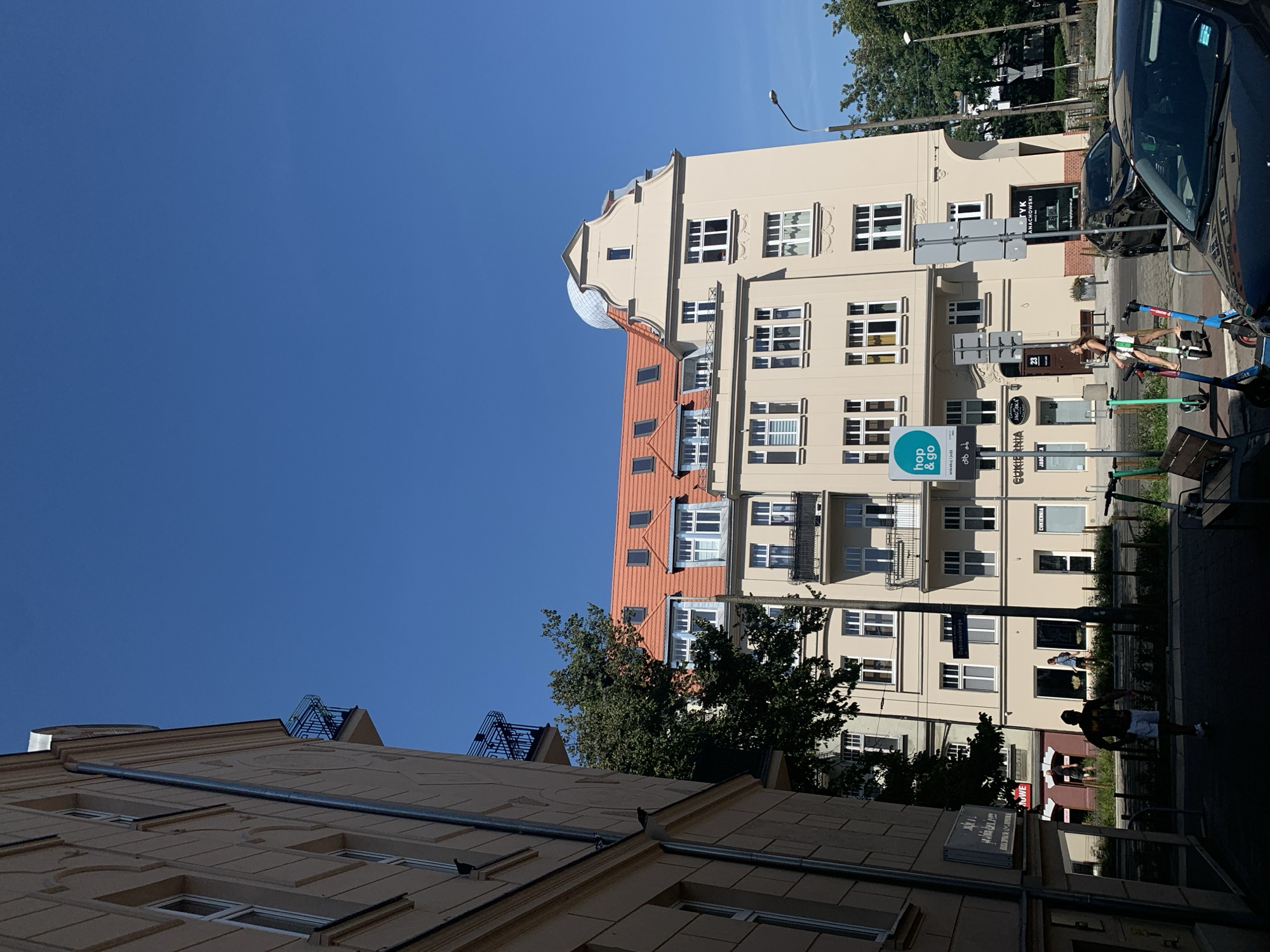 This is a daytime, eye-level shot of a street scene in Poznan, Poland. The image is dominated by a light-colored, multi-story building with a red tiled roof and dormer windows. In the foreground, a dark-colored car is parked on the right, and a person is walking on the left, silhouetted against the bright light. Several electric scooters are parked on the sidewalk.
The building's facade features numerous windows, some with balconies. A sign for "hop & go" is visible on the building, along with other signage in Polish that appears to be for shops and businesses. The street is paved, and trees line the background. The sky is a clear, bright blue.
The specific location is on Dąbrowskiego Street in Poznan, Poland, as indicated by a street sign in the image. The architecture suggests an older European city.
