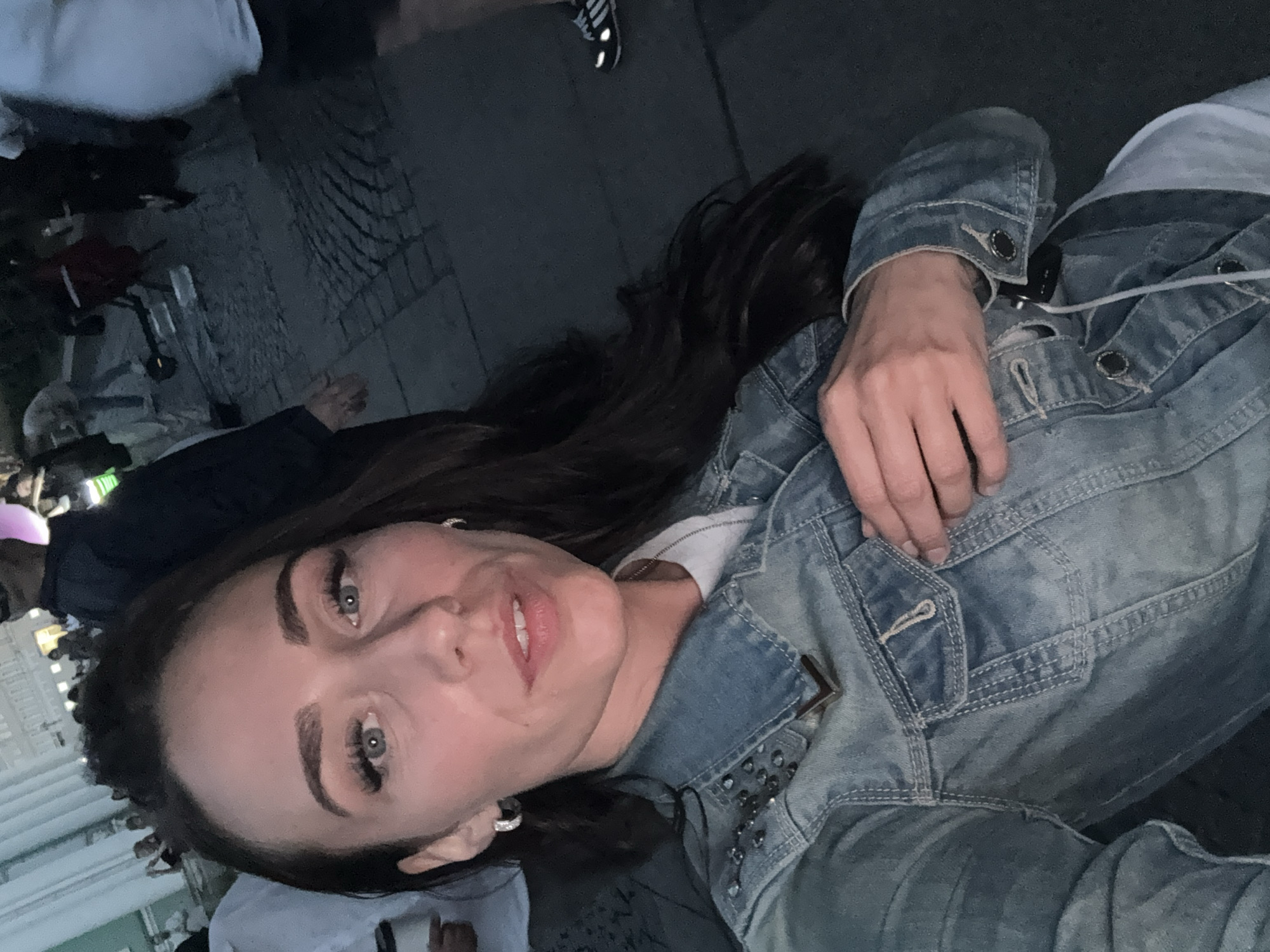 The image, rotated sideways, captures a close-up selfie or portrait of a young woman in a dimly lit outdoor urban environment, indicative of evening or night. She has long, dark hair, striking light-colored eyes, and wears a light blue denim jacket, possibly adorned with studs, over a white top. Her lips are slightly parted in a subtle smile, conveying a confident and relaxed demeanor. She also sports an earring.
The blurred background shows indistinct shapes of other individuals and ground paving, possibly cobblestones. A black shoe with white Adidas stripes is partially visible in the upper background. Hints of artificial lighting contribute to a lively, populated setting, likely a social gathering or event in Saint Petersburg, Russia. The overall atmosphere is informal and dynamic.