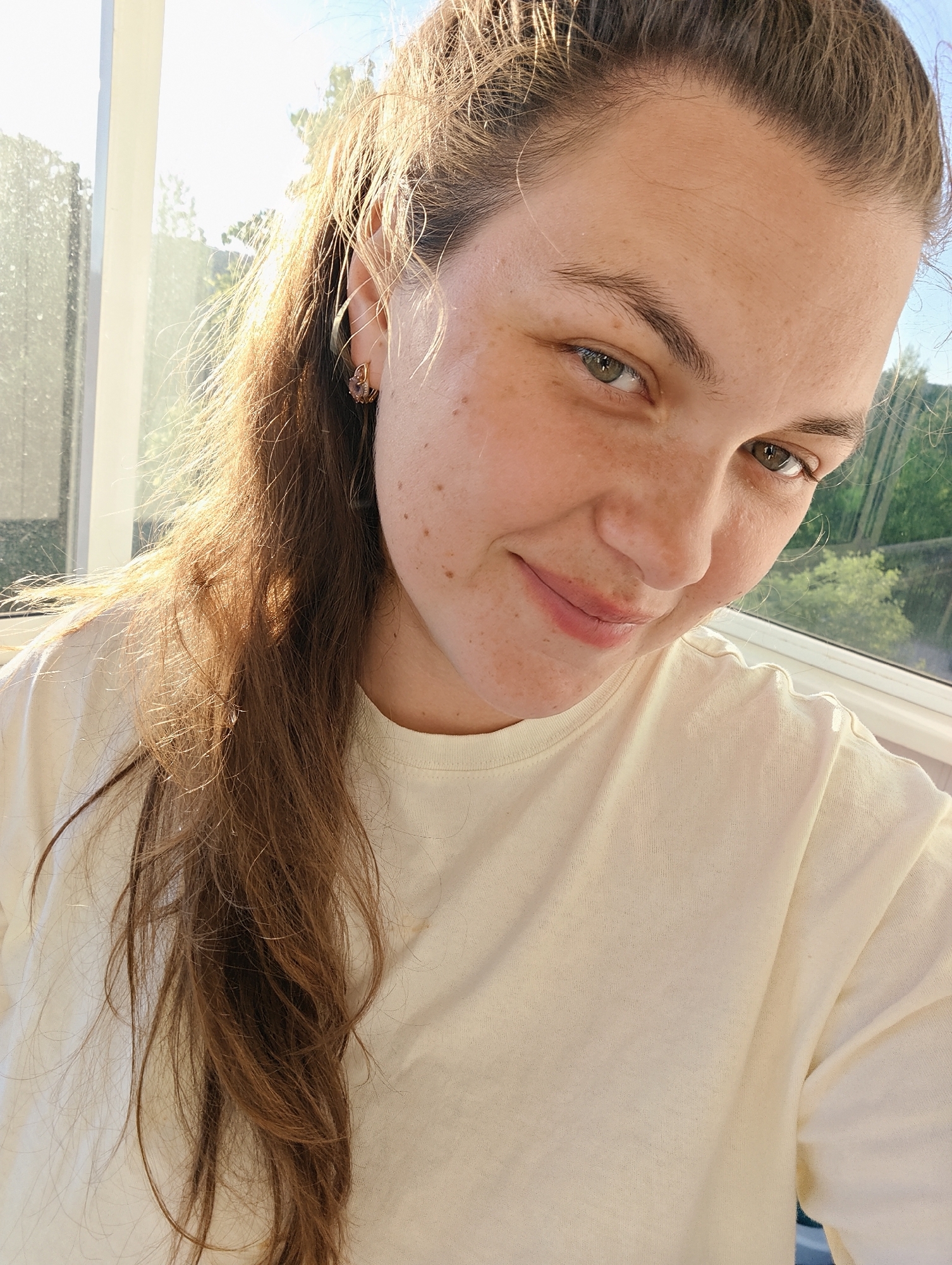 A brightly lit, close-up selfie-style portrait features a young woman with long, brown hair, partially pulled back, and loose strands framing her face. Her light skin is adorned with numerous freckles across her nose, cheeks, and forehead. She has green eyes and offers a gentle, closed-mouth smile, conveying a calm and friendly demeanor. She is wearing a simple, pale cream or light yellow t-shirt. A small, gold-toned earring with a reddish-purple accent is visible in her right ear.
Strong, warm natural sunlight streams from behind and to her left, creating a radiant halo around her hair and illuminating her features. The setting appears to be indoors, positioned next to a window. Through the window, a clear sky and lush green trees are visible, indicating an outdoor, natural environment. This bright lighting suggests it is daytime, likely morning or late afternoon, under sunny weather conditions in Chaikovsky, Russia. There is no visible text in the image.