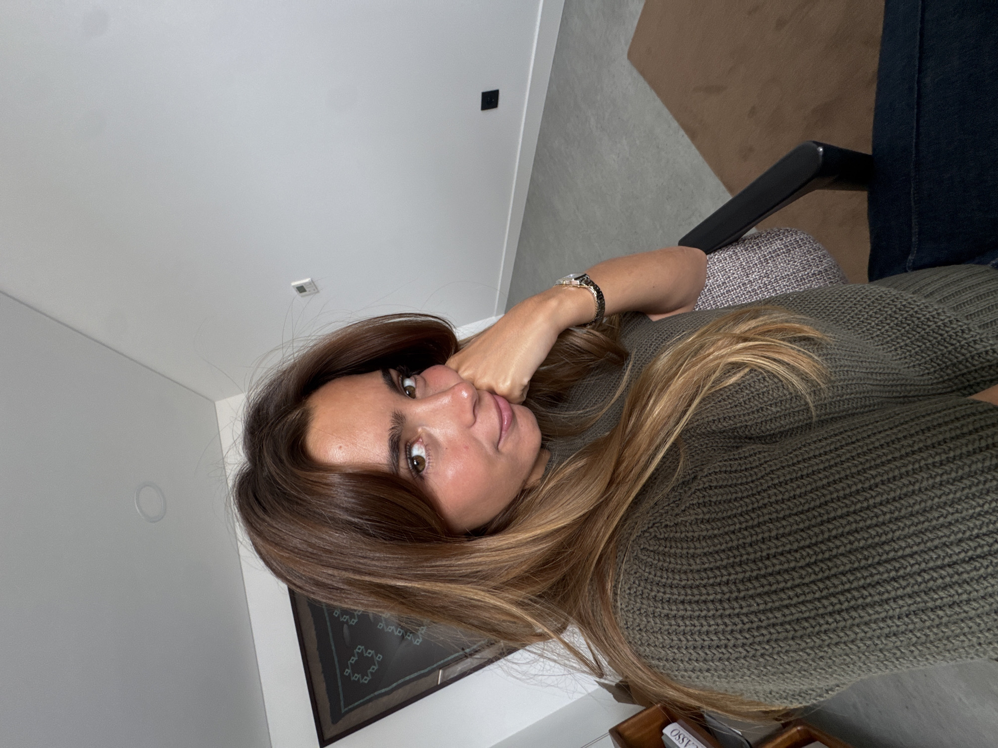 The image shows a woman reclining indoors, looking directly towards the camera with a calm expression. She has long, wavy brown hair with lighter highlights, and is wearing a dark olive green knitted sweater. Her left hand is raised to her chin, adorned with a watch featuring a dark band and a bracelet.
The setting appears to be a modern interior, possibly a living room. The background includes light-colored walls; one wall features a black square light switch or outlet, and another has a white circular fixture. Behind her, parts of a textured, light grey upholstered armchair or sofa, a dark armrest, and a dark blue fabric (potentially clothing or another furniture item) are visible, along with a brown rug. Below her, a framed artwork with dark geometric patterns can be partially seen. On a wooden surface in the bottom right corner, the partial text "CASSO" is visible, suggesting "PICASSO". The location context is Torres Vedras, Portugal.