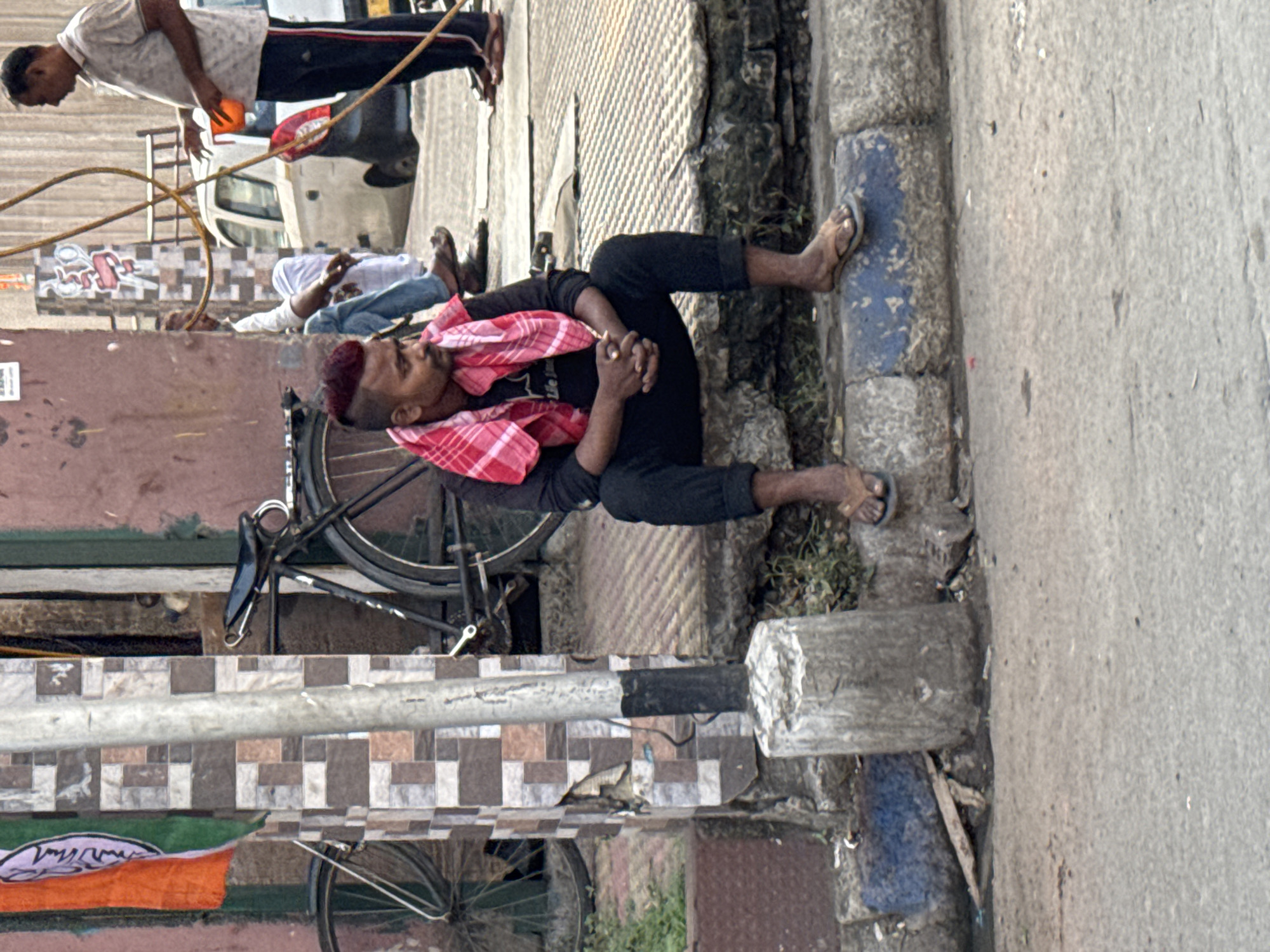 A man with a red-dyed mohawk is lying down on the side of a street in Kolkata, India, wearing black clothing and sandals. He is using a red and white checkered cloth as a pillow, his hands clasped. A bicycle is parked next to him, partially obscured by a pole decorated with brown tiles and a flag bearing an orange and purple symbol. In the background, other men are reclining on the ground near a parked white van. One man is standing nearby, holding an orange object in his hand and standing by some yellow cables.
