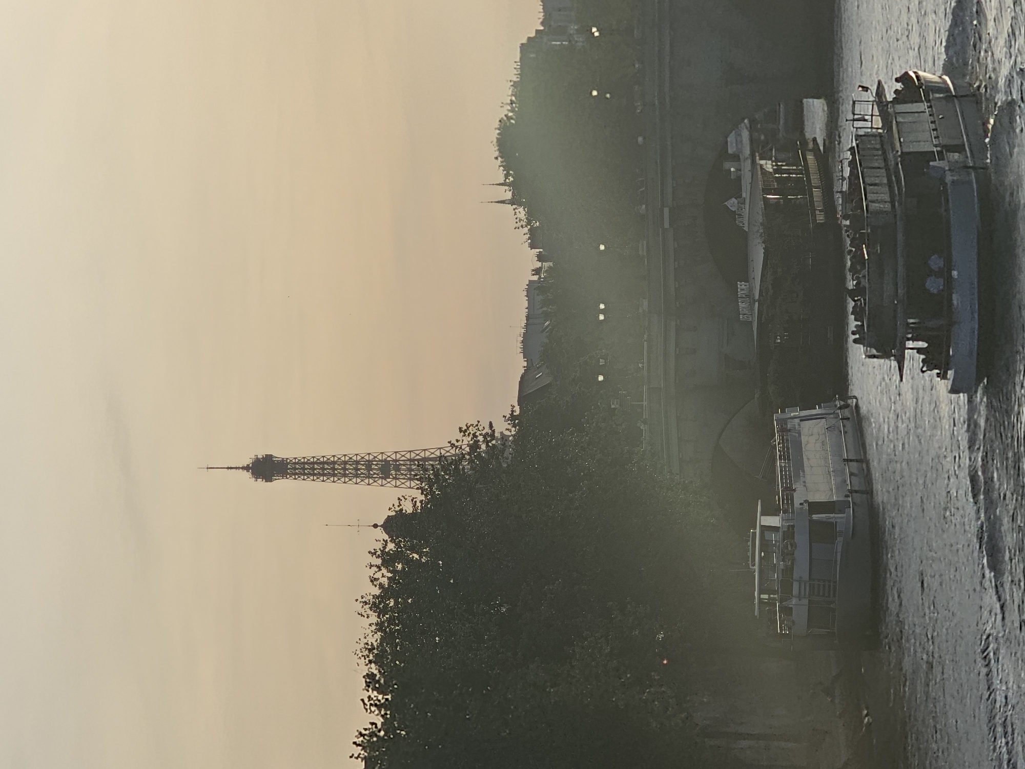 The image displays a serene, horizontal view of Paris, France, captured during the golden hour, likely sunrise or sunset, given the warm, pale yellowish-orange sky that dominates the upper portion of the frame.
In the mid-ground, the iconic Eiffel Tower stands prominently against the soft sky, its distinctive lattice ironwork clearly visible. Below and to the left of the tower, a dense canopy of dark green trees fills much of the middle and lower-left section of the image, partially obscuring the tower's base and the urban landscape behind them. Beyond the trees, the silhouette of distant buildings and a noticeable church spire can be discerned against the horizon.
To the right, a calm body of water, indicative of the River Seine, stretches across the foreground and mid-ground. Two large passenger boats are visible on the water. The boat further away from the viewer appears to be a multi-deck vessel, and the partial text "BATEAUX MOUCHES" is discernible on its side, identifying it as a common tourist river cruise. Indistinguishable figures, likely passengers, can be vaguely seen on its deck. Another boat, closer to the viewer, is also a multi-deck design with railings and windows. The scene evokes a peaceful and classic Parisian vista.