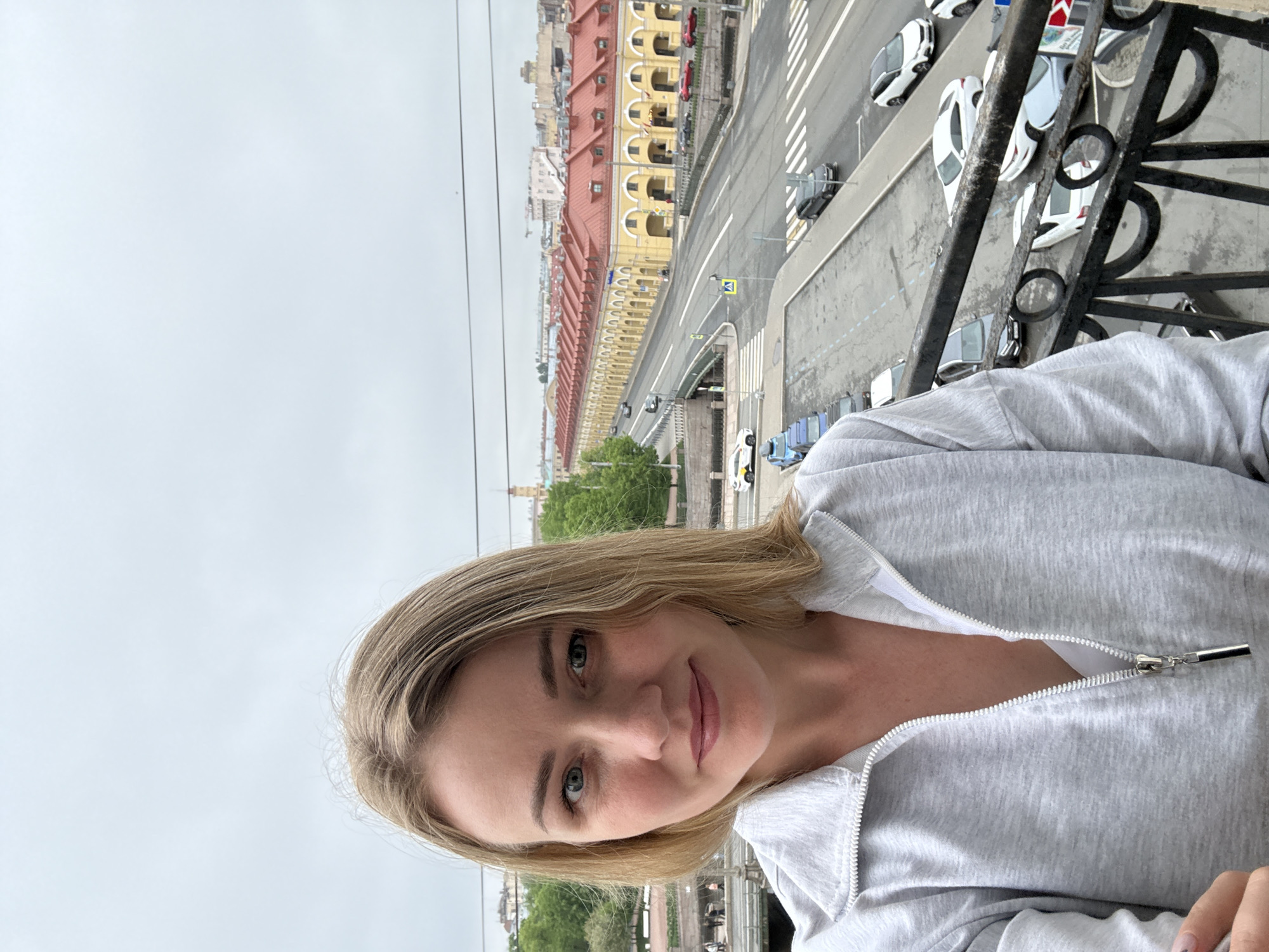 A woman with blonde hair and blue eyes is wearing a gray zip-up jacket. She is looking at the camera and smiling slightly. She is standing on a balcony with a black wrought iron railing. Behind her is a street in Saint Petersburg, Russia. Cars are parked along the street, and a long yellow building with a red roof is visible in the background. The sky is overcast.