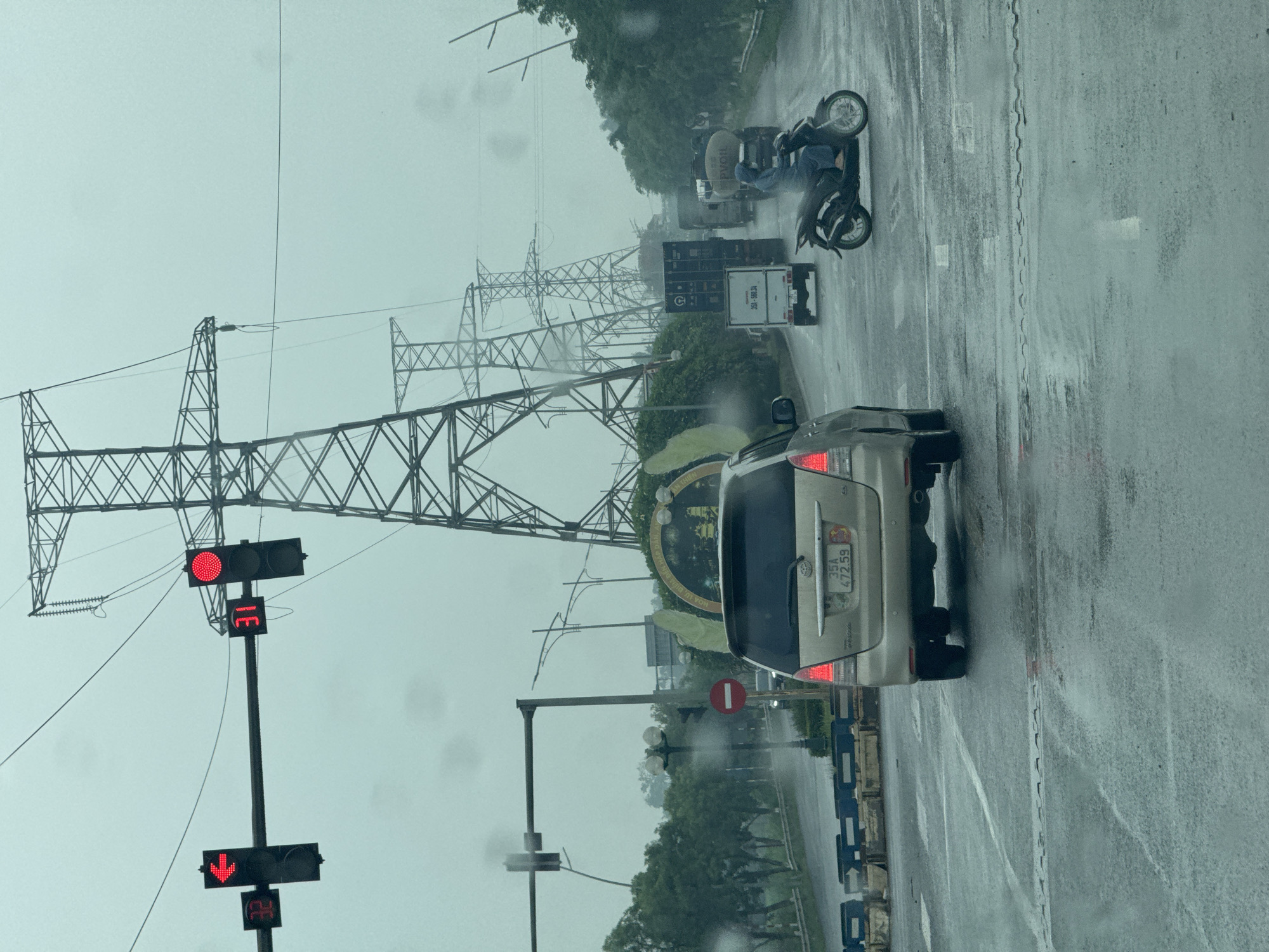 A wet, overcast day in the city of Hoa Lư Ward, Vietnam, shows a traffic intersection. In the foreground, a beige Toyota Innova is stopped at a red light. The license plate reads "35A 472.59". To the left of the Innova, a traffic light shows a red left-arrow signal and a red circular signal. Next to it, a digital timer displays "32". Further down the road, a motorcycle with a rider wearing a blue rain suit is moving through the intersection. Several trucks, including a container truck and a tanker truck, are also visible. Tall electrical transmission towers are seen lining the road. The road surface is wet, indicating recent or ongoing rain, with visible puddles and reflections. A large green archway with lettering is partially visible in the background, suggesting a formal entrance or landmark.