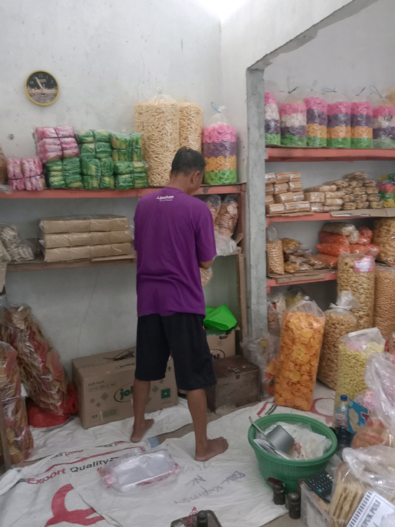 The image shows a small shop in Sugihwaras, Indonesia, filled with shelves of packaged snacks. A man wearing a purple t-shirt and black shorts is standing in the middle of the shop, facing away from the camera. He appears to be sorting or organizing the snacks. The shelves are packed with various types of crackers, chips, and other snacks in colorful packaging. There are also some boxes and plastic bags on the floor. The overall impression is of a well-stocked snack shop, possibly a small family-run business.