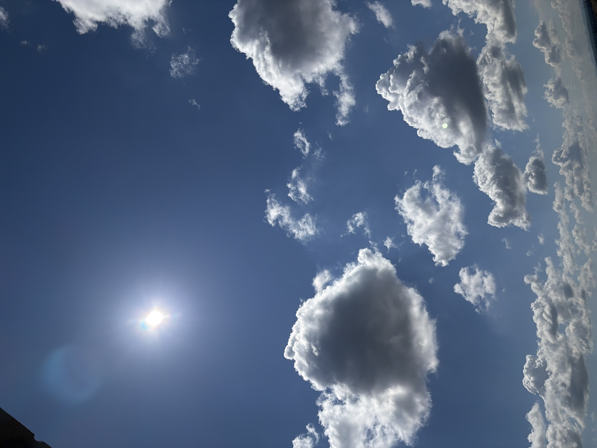 The image captures a daytime view of the sky in an unknown city. The sky is predominantly blue, with scattered cumulus clouds of varying sizes and shapes. The sun is visible in the lower left corner, emitting a bright glare.