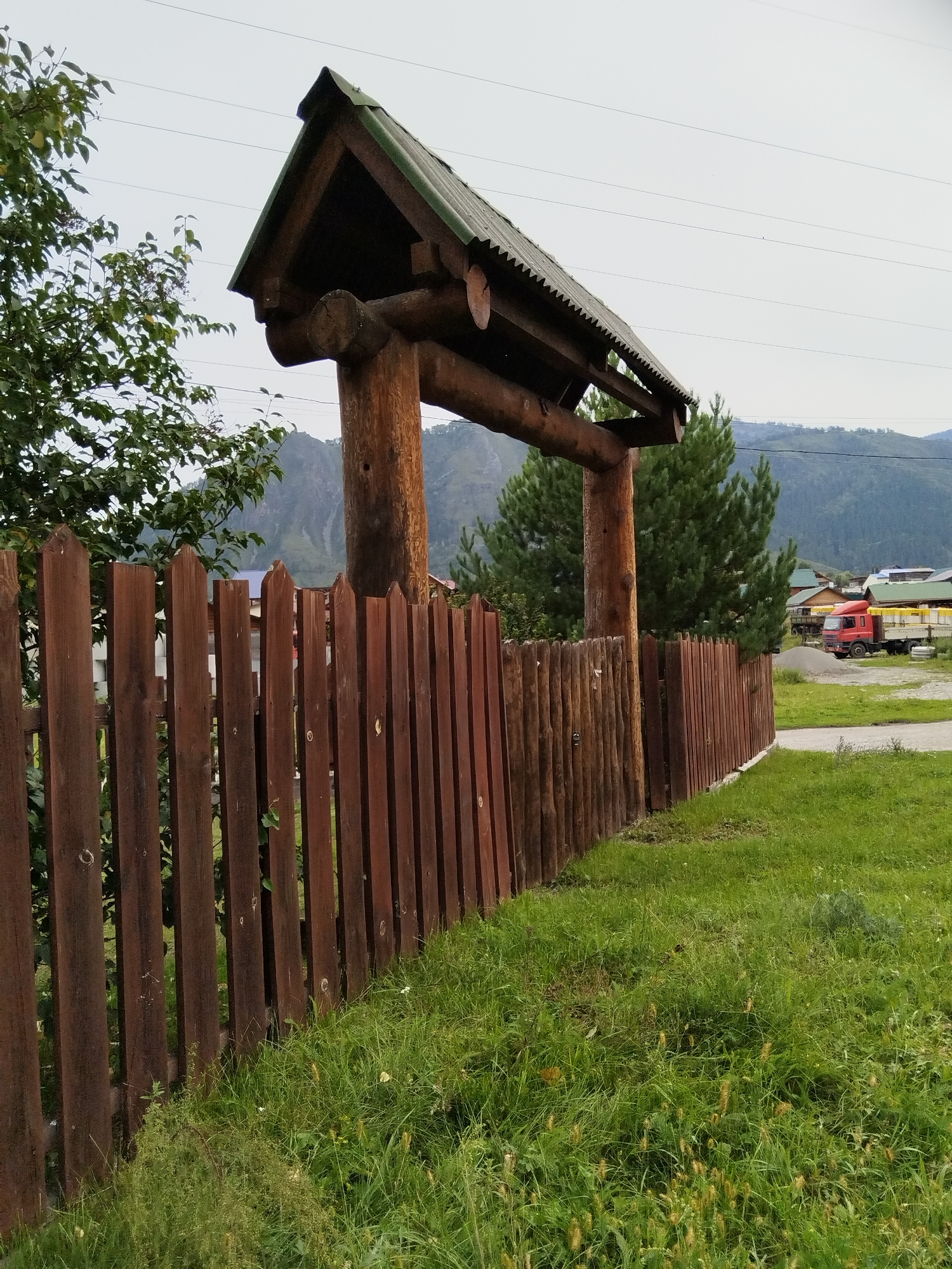The image depicts a wooden archway constructed from logs, topped with a dark, weathered roof featuring a serrated edge. This archway stands behind a rustic wooden fence made of vertical, dark brown planks, which extends out of frame to the right. Lush green grass covers the foreground and surrounds the fence. In the background, pine trees are visible, along with a cluster of houses with green roofs, suggesting a village setting. Further back, rolling hills or mountains create a natural backdrop under an overcast sky. A red truck with a yellow cargo is parked on a dirt area to the right of the fence. The scene appears to be located in Chemal, Russia, a town known for its natural beauty and wooden architecture.