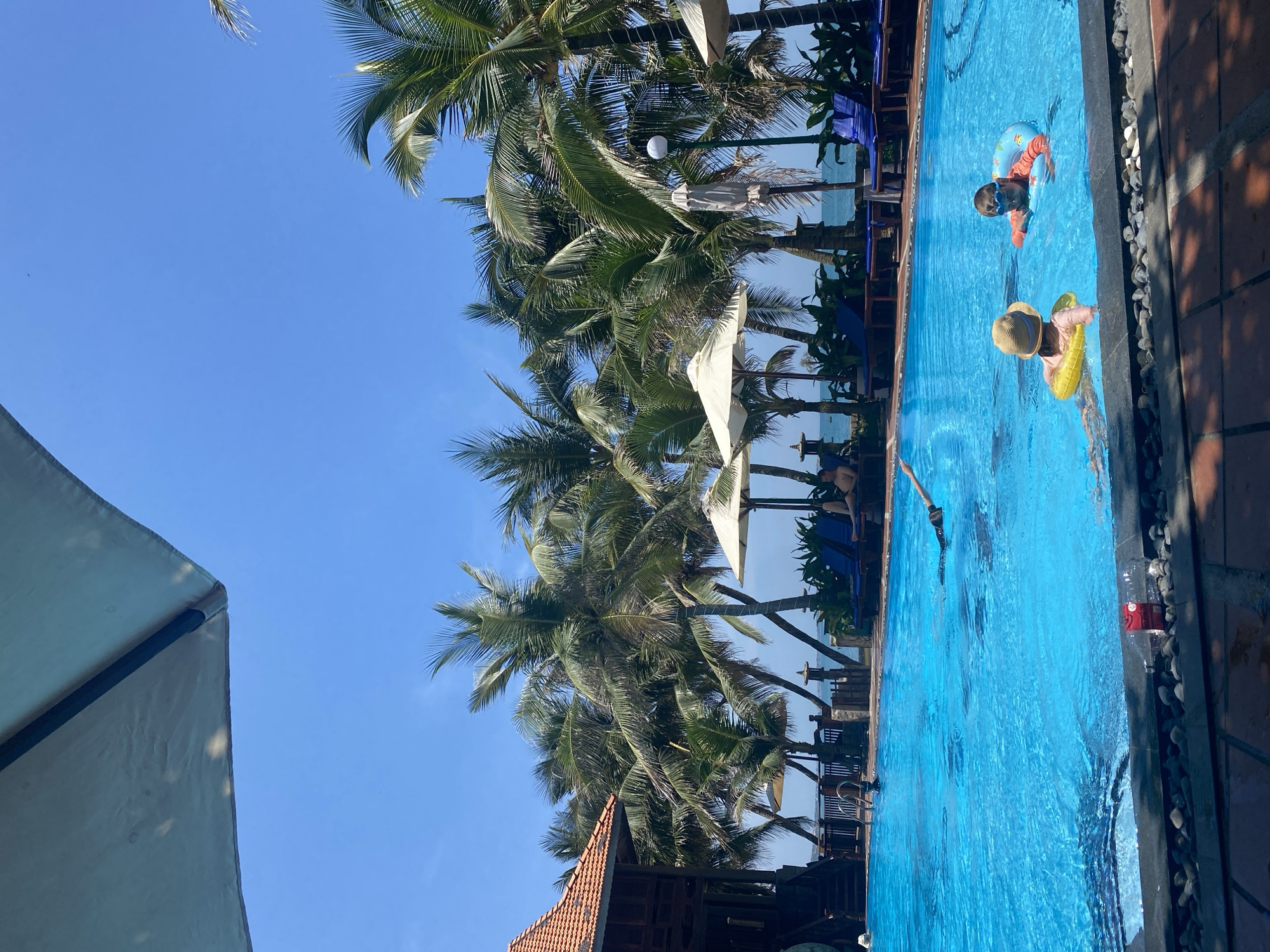 The image depicts a bright, sunny day at a resort pool, likely in Phu Thuy Ward, Vietnam, judging by the abundance of palm trees and the architectural style of the building. In the foreground, a vibrant blue swimming pool dominates the scene. Two children are visible in the pool: one wearing a straw hat and a yellow floatation ring, and another wearing goggles and an orange floatation ring. A person is swimming in the mid-ground of the pool, with their body partially submerged and arms outstretched. In the background, lush green palm trees line the edge of the pool, leading towards a view of the ocean under a clear blue sky. Beach chairs and umbrellas are set up near the pool, some occupied by individuals relaxing. A multi-story wooden building with an orange tiled roof is visible to the left, adding to the tropical resort ambiance. The poolside is edged with dark stones and reddish-brown paving.