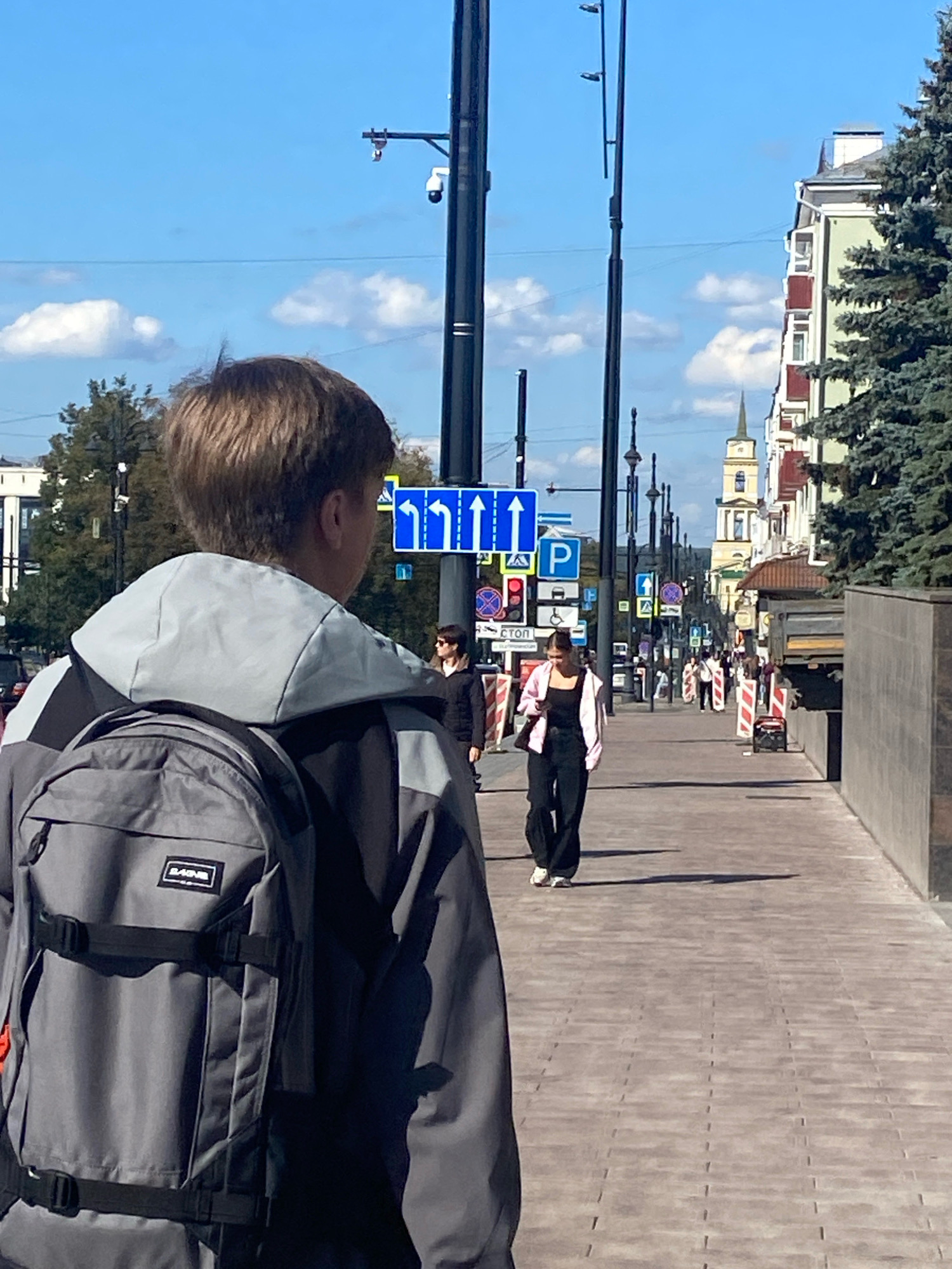 This is a street-level, medium-distance, eye-level shot in Perm, Russia, showing a young person from behind, wearing a gray jacket and backpack, walking on a paved sidewalk. The person is facing away from the camera and looking down a street lined with buildings and trees. The sky is blue with some clouds. In the background, other pedestrians are visible, as well as street signs and traffic lights. The street appears to be a busy urban area with architectural styles suggesting pre-Soviet and post-Soviet construction.