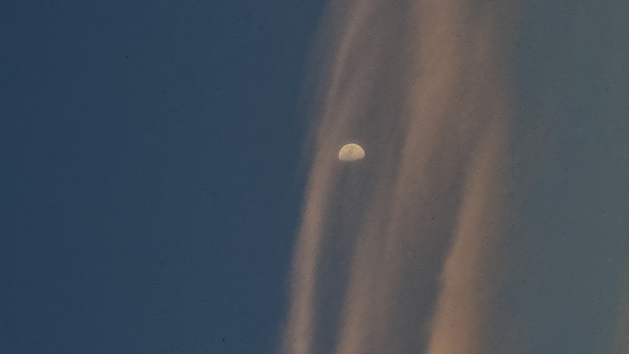 A crescent moon is visible in the sky, appearing as a pale yellow arc against a deep blue background. The moon is partially obscured by wispy, vertical cloud formations that are tinged with a peachy-pink hue, suggesting either sunset or sunrise. The clouds extend diagonally across the frame from the right side towards the center. The overall scene depicts an atmospheric celestial view, likely captured in the evening or early morning. No people or explicit objects are present, and there is no visible text. The setting appears to be an outdoor, open environment given the celestial display.