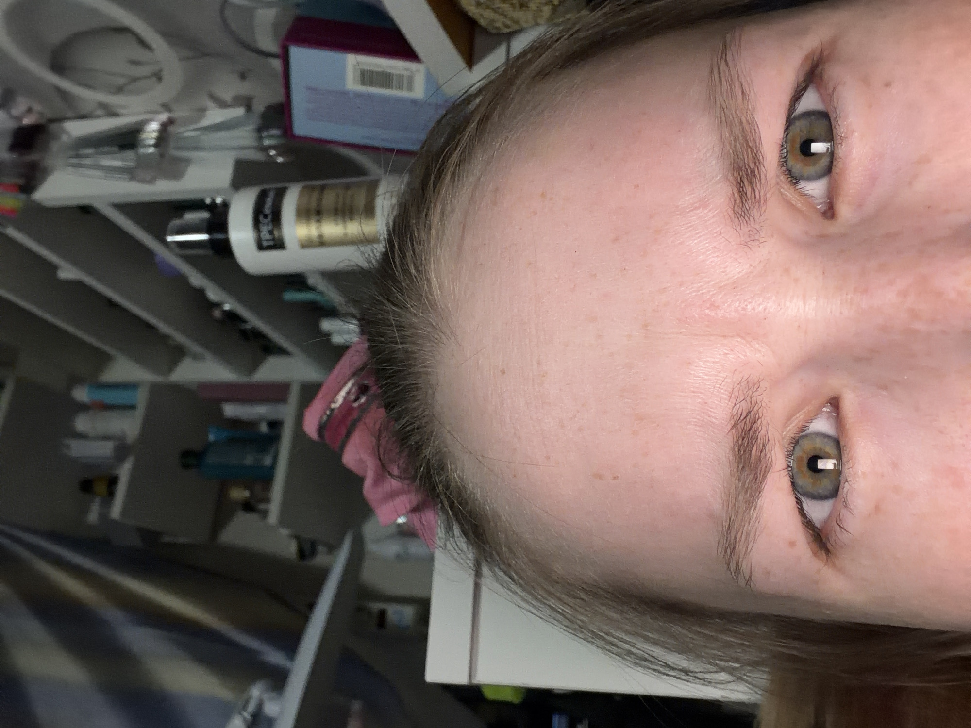 A close-up portrait shows a person's face from the forehead to the lower part of the nose. The person has light skin, freckles, and grey eyes. They are surrounded by a bathroom with a shelf, various beauty products, and a pink scrunchie. The image does not show anything that allows identifying the location as Berezniki, Russia.