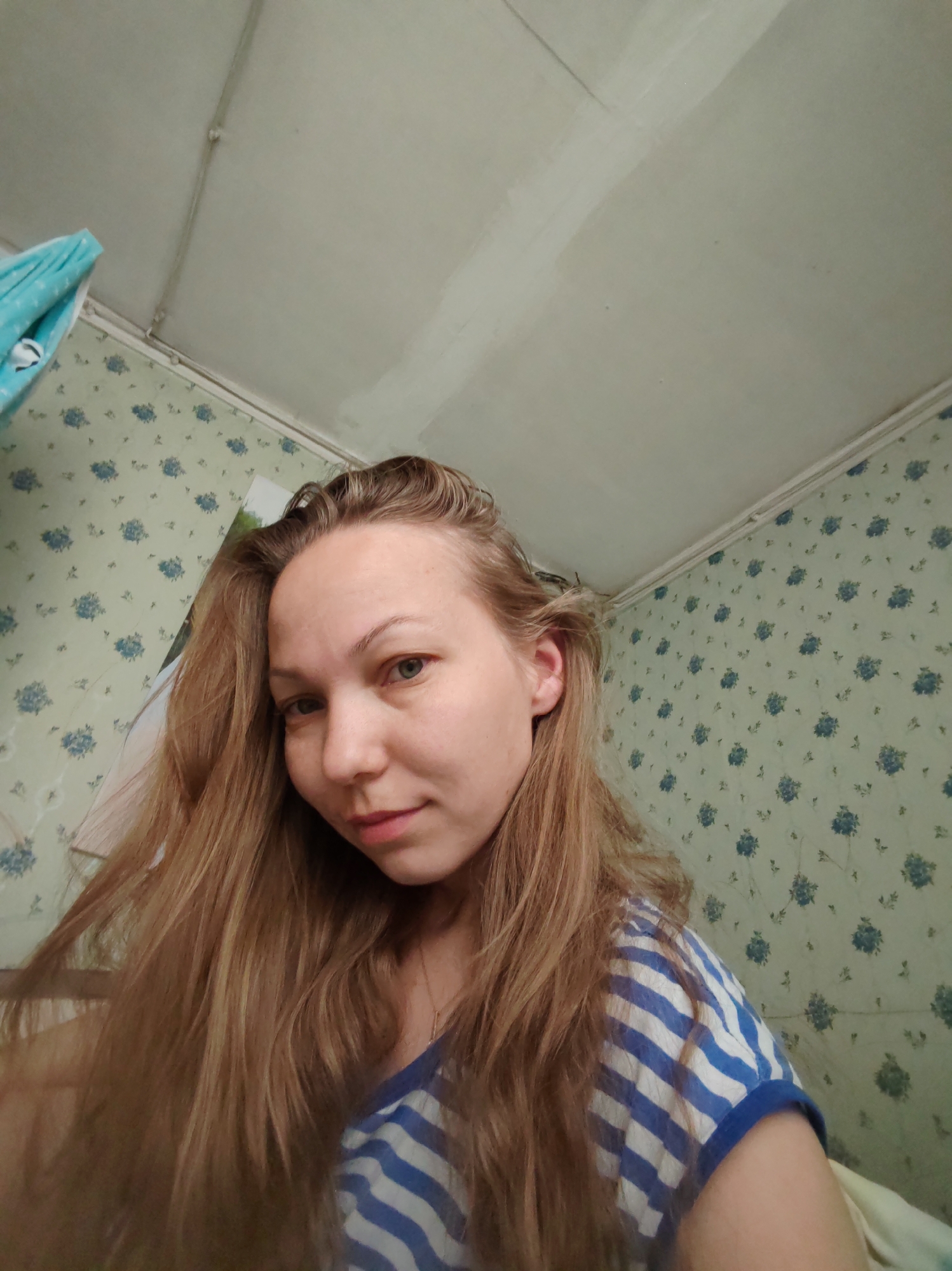 A person with long, light brown hair and fair skin is facing the camera. She is wearing a blue and white striped shirt. The background includes a ceiling with visible electrical wiring and walls covered in a light green wallpaper with a pattern of small blue flowers. A blue towel is hanging in the upper left corner of the frame. The location appears to be an interior space in a building in Чухлэм, Russia, given the context.