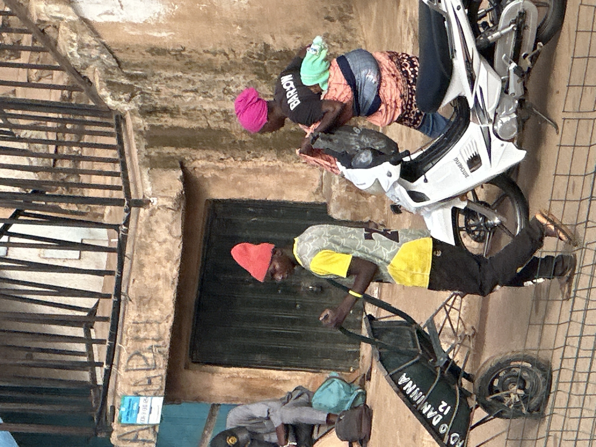 In Bukuru, Nigeria, a street scene unfolds. A young man, wearing a bright red beanie and a yellow and gray patterned shirt with the number "2" visible, pushes a dark green wheelbarrow. The wheelbarrow has white lettering on its side, partially obscured, but "DANINNA" and "12" are legible. To his right, a woman, adorned in a vibrant pink headwrap and a black t-shirt with "BARION" printed on the back, carries a baby wrapped in green and orange fabric on her back. She is also holding a bundle of fabric in her arms. Beside them, a white motorcycle with black accents stands parked. In the background, a man in a gray outfit and a cap sits on the ground near some bags. The setting appears to be an urban environment, with a weathered building featuring a dark doorway behind the individuals. A metal railing is visible above, casting shadows. The ground is a dusty, uneven surface, and a wire fence runs across the foreground.