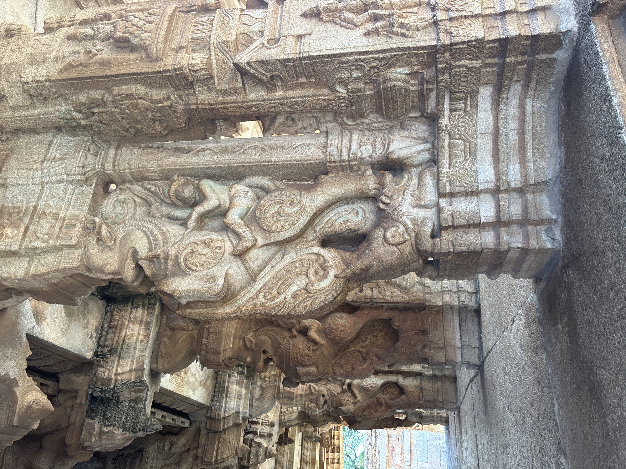 This image shows detailed stone carvings in Hampi, India. The carvings depict figures on mythical creatures, potentially lions or similar beasts. The stone has a weathered appearance, indicative of age and exposure to the elements. The architecture suggests a temple or similar historical structure, with rows of carved supports creating a corridor or passageway. Light filters through the opening at the end of the corridor, indicating an outdoor setting. The scale and complexity of the carvings suggest skilled craftsmanship and significant cultural importance.