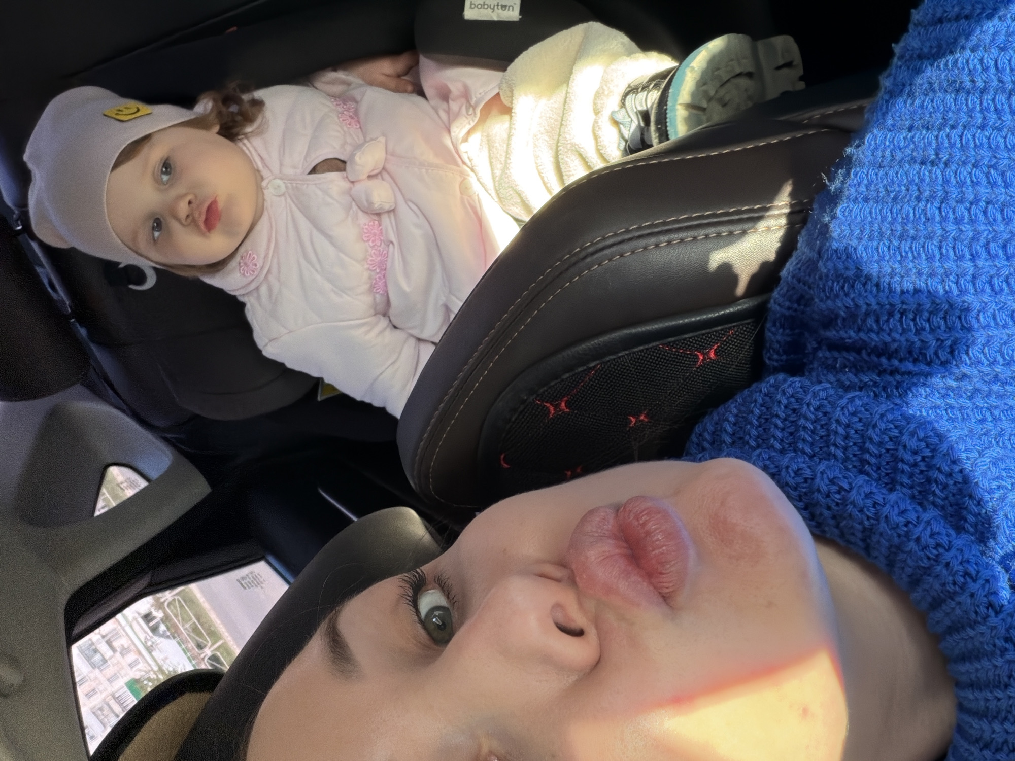 The image depicts a woman and a young child inside a car. The woman, wearing a blue sweater, is in the foreground, with the lower half of her face visible. She is looking up towards the camera with a pursed lip expression. Behind her, in a car seat, is a child wearing a light pink jacket and hat with a smiley face emblem. Another item of clothing, light-colored, is also in the car seat. The car seat itself is black leather with red detailing. Outside the car window, the environment indicates a building, potentially in Chaikovsky, Russia.