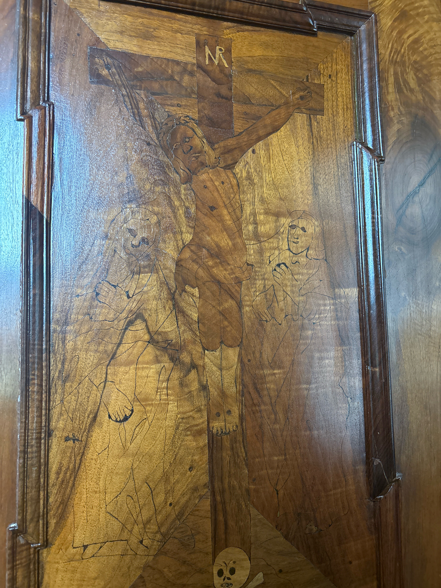 The image shows a close-up view of a wooden intarsia panel, likely a cabinet door, depicting a scene of the Crucifixion of Jesus Christ. The artwork is rendered in shades of wood, with darker wood for the figures and outlines against a lighter background. Jesus is shown on the cross, with the letters "INRI" above his head. To his left and right are two figures, likely Mary and John the Evangelist, depicted with flowing robes and somber expressions. At the base of the cross, a skull and crossbones are visible, a memento mori symbol. The wood grain of the panel creates a textured backdrop. The framing around the panel appears to be dark, carved wood. This intarsia panel is part of a collection of decorative arts housed in the Hermitage Museum in Saint Petersburg, Russia.