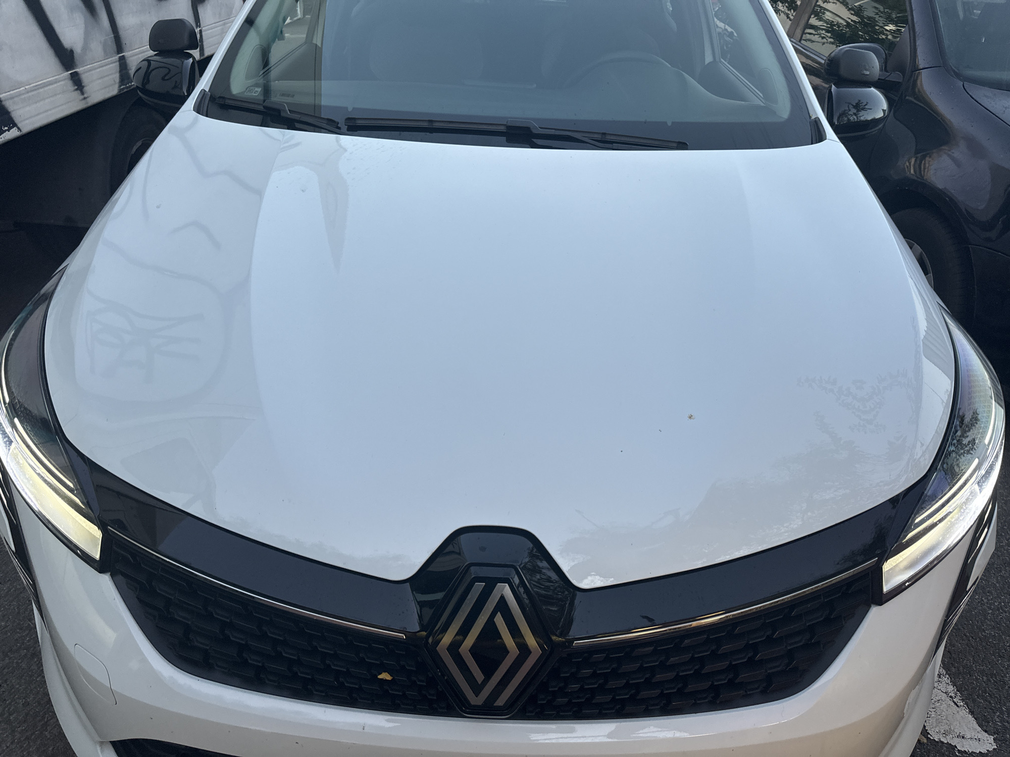The image shows the front of a white car, a Renault, parked on an asphalt surface. The car has a black grille with the Renault logo in the center and headlights on either side. In the background, to the left, is a portion of a white truck with graffiti on it. To the right of the car, another dark-colored car is parked. The scene appears to be outdoors in a city environment, possibly a street or parking lot. The lighting suggests it is daytime. There are no people visible in the image. Without further context, it is not possible to determine the specific location within the city of Murcia, Spain.