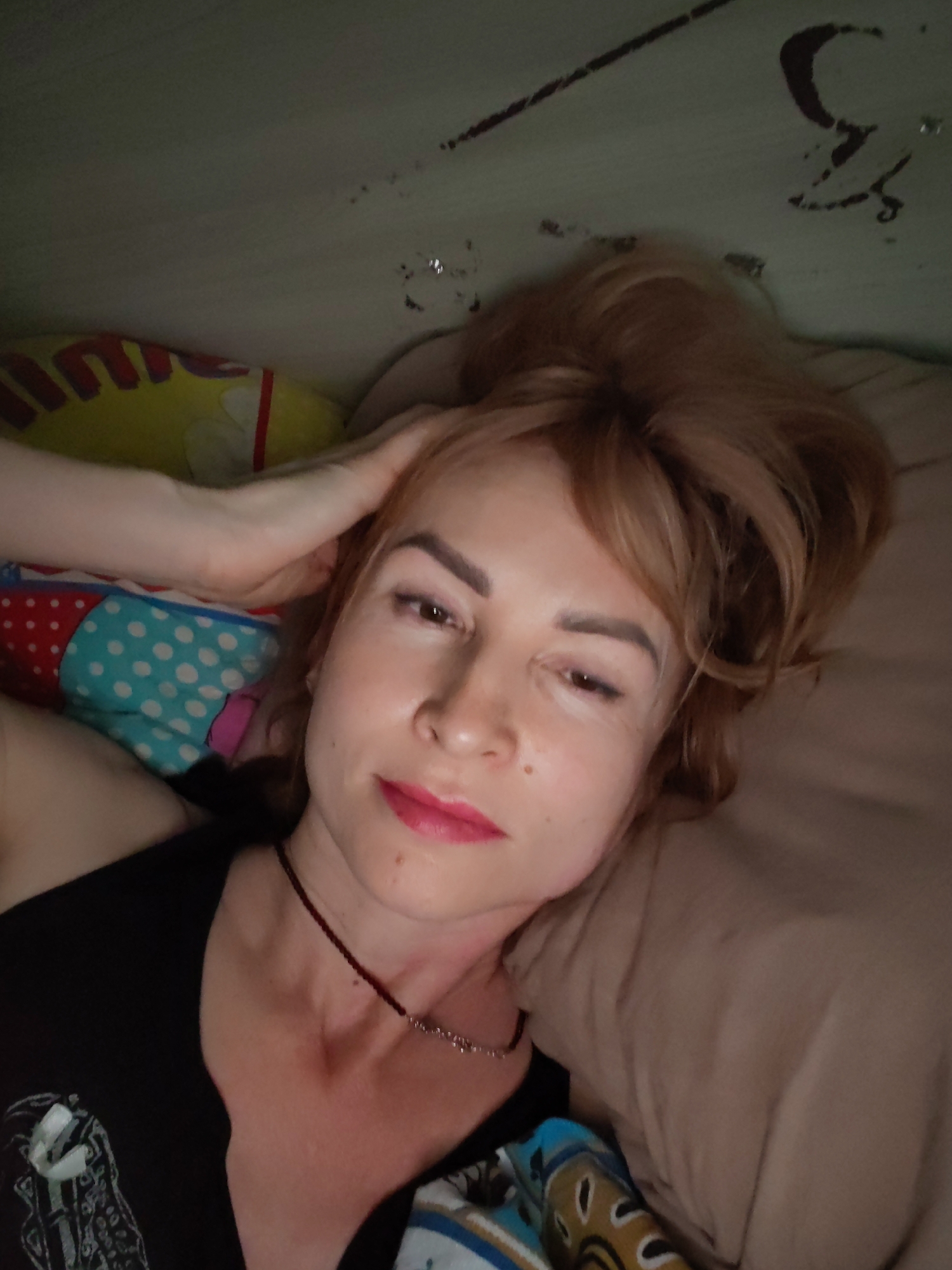 A woman with fair skin and light brown, tousled hair is lying down, possibly on a bed or resting area. Her head is tilted towards the camera. She is wearing a black top and a delicate necklace. Her left hand rests on her head. The background shows bedding with various patterns and colours. There is a wall behind the bed. The lighting appears to be somewhat dim. There is no indication of the city of Chaikovsky, Russia, in the image.