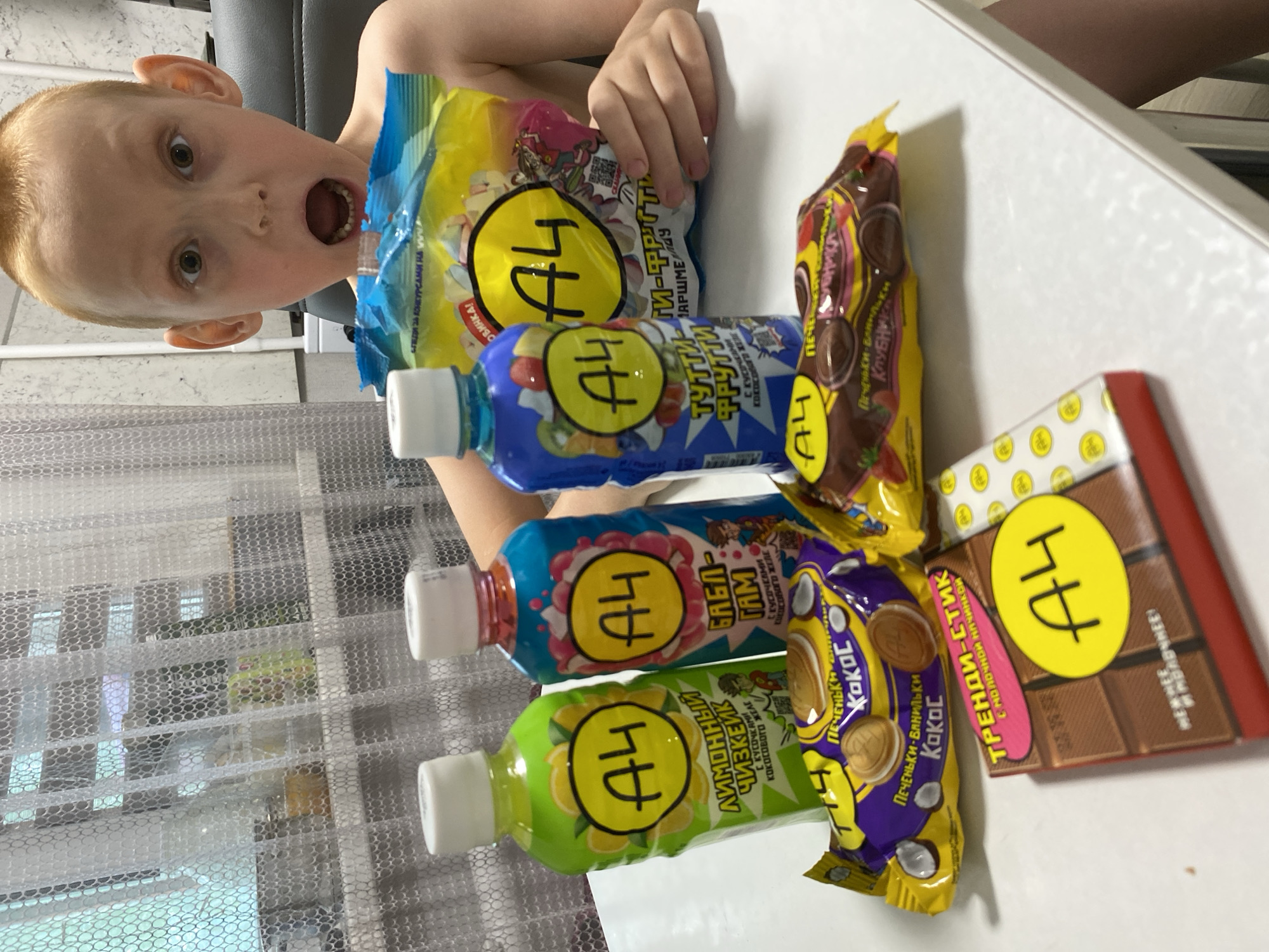 A young child, likely a boy with light hair, is captured mid-reaction, looking directly at the camera with wide, surprised eyes and an open mouth, expressing excitement or astonishment. He appears to be shirtless or wearing very light clothing.
The scene is an indoor setting, possibly a home in Noviy, Russia, where an assortment of "A4" branded food and drink products is displayed on a white table. The child is positioned behind these items, suggesting he is interacting with or about to enjoy them.
The products prominently feature the "A4" logo and include:
- A yellow bag of "ТУТТИ-ФРУТТИ МАРШМЕЛЛОУ" (Tutti-Frutti Marshmallows), labeled "НОВИНКА!" (New!).
- Three bottled drinks with coconut jelly pieces: a blue "ТУТТИ-ФРУТТИ", a pink "БАБЛ ГАМ" (Bubble Gum), and a green "ЛИМОННЫЙ ЧИЗКЕЙК" (Lemon Cheesecake).
- A purple and yellow bag of "ПЕЧЕНЬКИ-ВАНИЛЬКИ КОКОС" (Cookies-Vanilla Coconut).
- A red and brown "ТРЕНДИ-СТИК" (Trendy Stick) chocolate bar with milk filling, described as "МЕЛКИЙ ШОКОЛАД" (Small Chocolate).
In the background, a translucent white mesh curtain or blind covers a window, indicating it is daytime. The child's exaggerated facial expression is a notable detail, conveying strong emotion in response to the colorful array of snacks. The products themselves, with their bright packaging and specific flavors, are clearly targeted towards a younger audience, aligning with the popularity of the "A4" brand often associated with a Russian YouTuber.