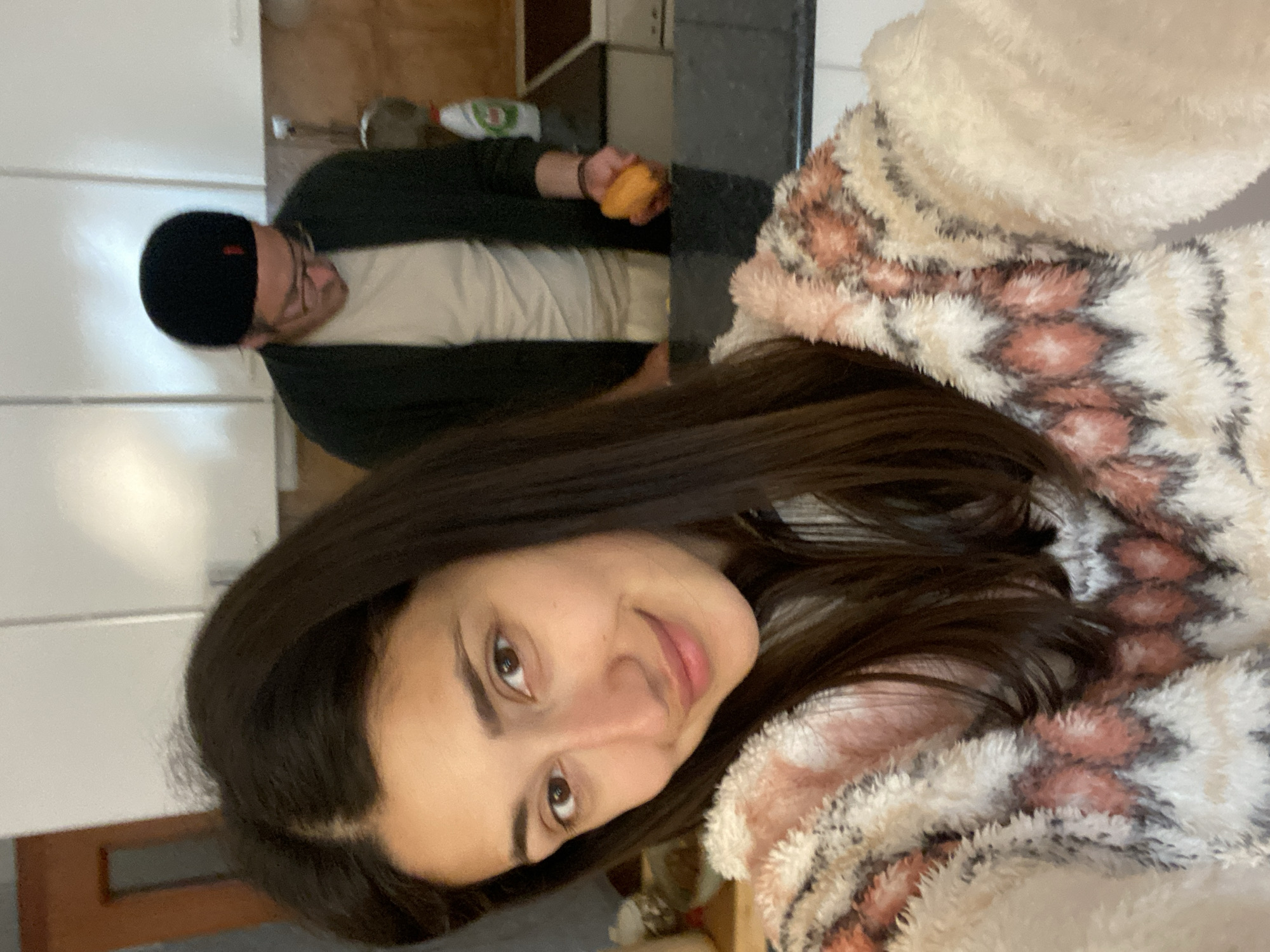 The image displays an indoor scene, indicated as being located in Sutomore, Montenegro, depicting two individuals in what appears to be a residential kitchen. In the immediate foreground, an adult female with long, dark brown hair and a light complexion faces the camera, holding a device consistent with a smartphone to capture a selfie. She exhibits a slight smile and is dressed in a fluffy garment featuring a pattern of white, beige, and reddish-pink geometric designs. A dark, possibly black, countertop is visible below her shoulder level, acting as a partition within the frame.
In the background, positioned behind the countertop, an adult male is observed leaning forward. He wears a black beanie with a small red detail, round-rimmed glasses, a light-colored t-shirt or undershirt, and a dark-colored open cardigan or jacket. His attention is directed downwards towards an orange fruit or object held in his right hand. The background environment consists of white upper kitchen cabinets, a light-tiled backsplash, and a continuation of the dark countertop. On this counter, a white bottle with a green cap and a red label, identifiable as Fairy brand dish soap, is clearly visible. To the man's right, a dark surface resembling a stovetop or sink is present. A portion of a wooden door frame is also discernible in the upper left background of the scene.