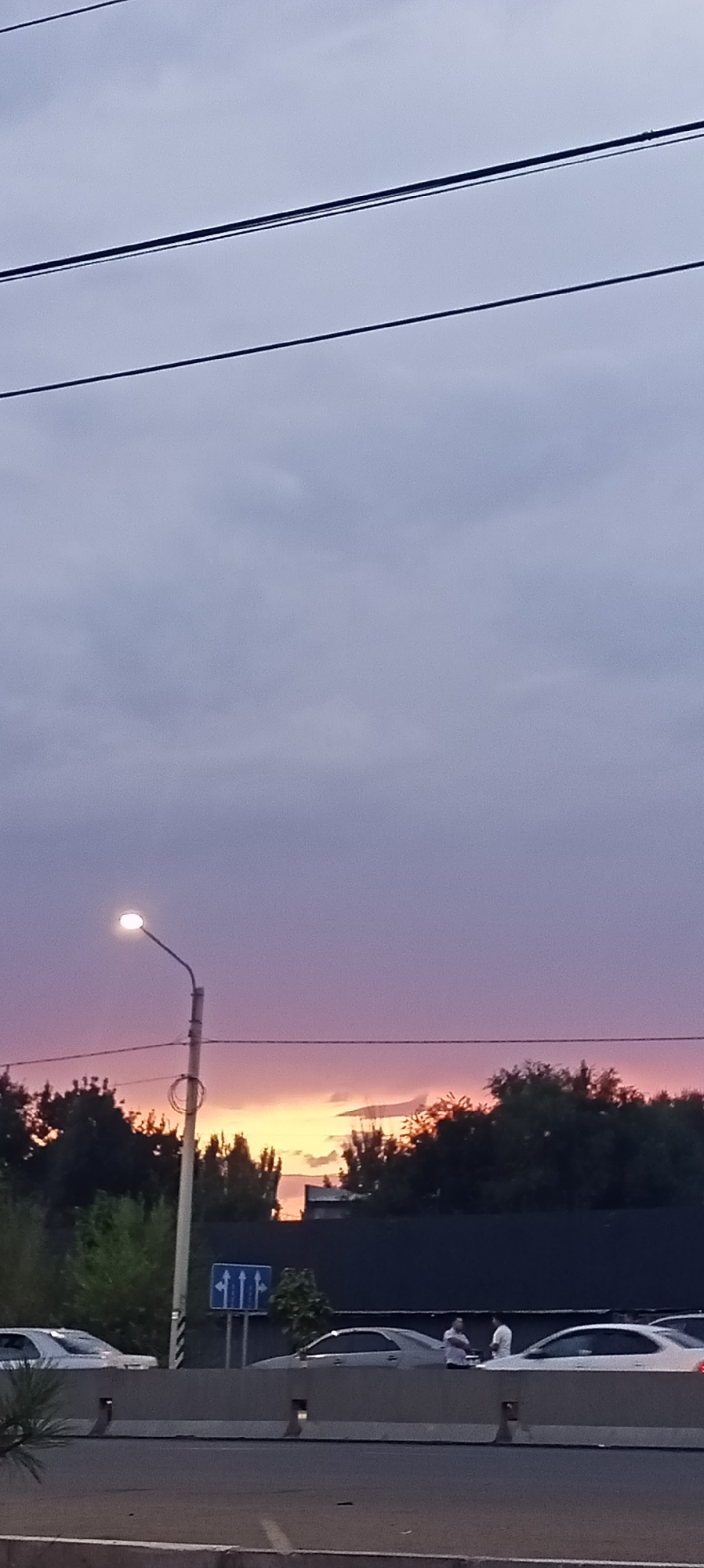 A sunset is depicted in Sokuluk, Kyrgyzstan, with clouds colored in shades of purple and orange. In the foreground, a street light illuminates the scene. Multiple cars are parked on the side of the road, and a few people are standing near them. Power lines are visible in the sky above.