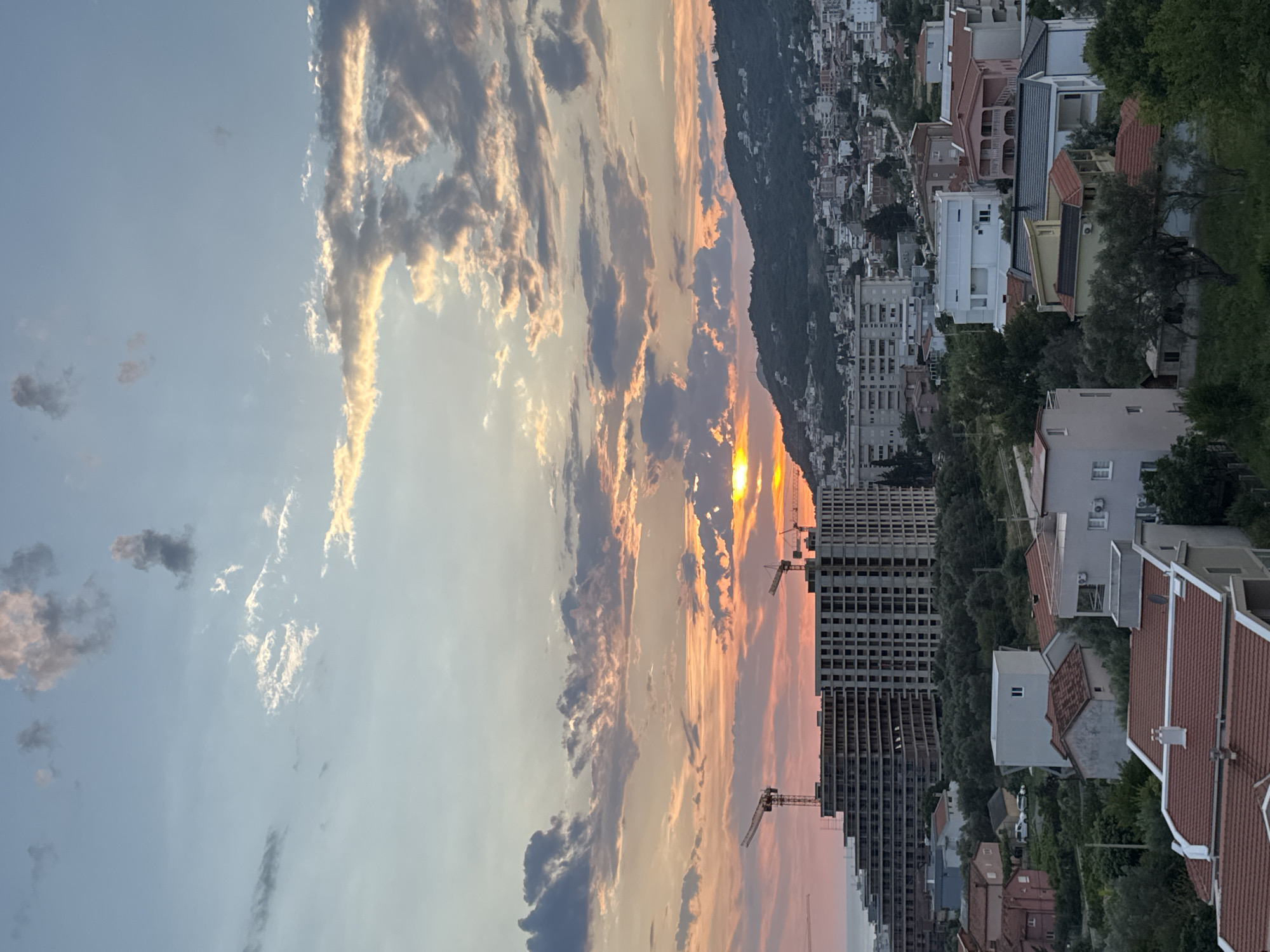 The image captures a sunset scene over the city of Šušanj, Montenegro. The sky is a gradient of blues and oranges, with scattered clouds illuminated by the setting sun. A large, unfinished building complex dominates the mid-ground, with several construction cranes visible. Below this complex, a cluster of residential buildings with red-tiled roofs is nestled amongst lush green trees. Further in the background, a hillside covered in more buildings and vegetation slopes upwards. The overall impression is one of urban development set against a natural, scenic backdrop.