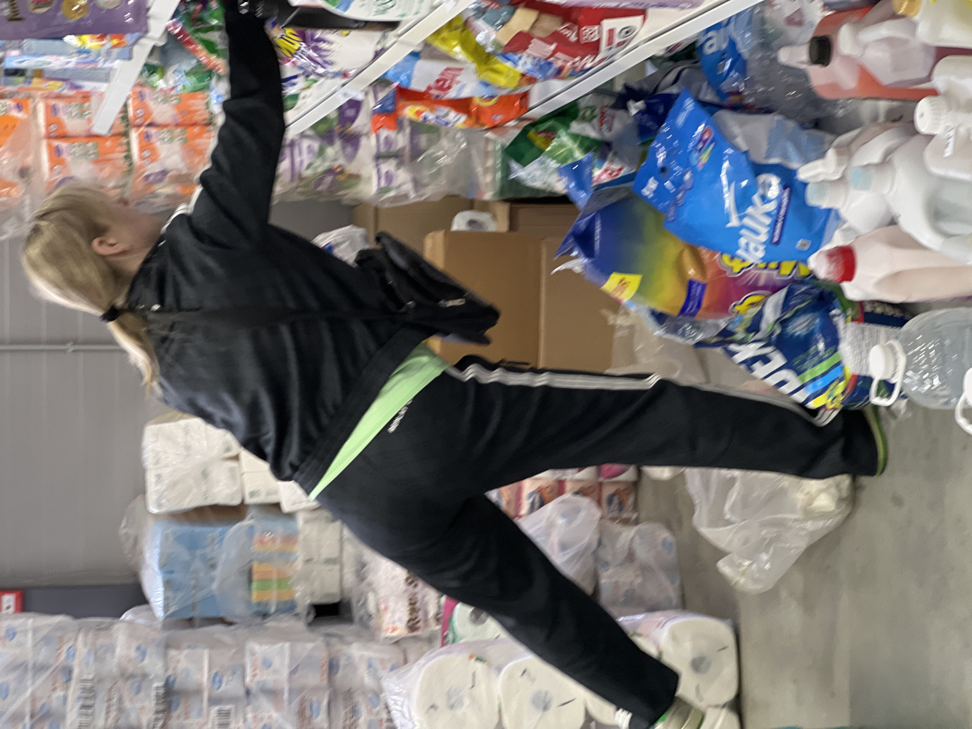 The image, taken in Krasnodar, Russia, captures a woman reaching for items on a high shelf or stacked goods in what appears to be a warehouse, storage area, or bulk shopping environment. The photograph is oriented sideways, depicting the woman stretching upwards with her right arm extended.
The subject is a woman with blonde hair, pulled back in a ponytail. She is dressed in a black tracksuit featuring lime green stripes along the sides of her pants, where a "Reebok" logo is faintly visible. She wears light-colored athletic shoes and has a black shoulder bag.
The scene is cluttered with various household products, primarily cleaning supplies and paper goods, stacked on shelves and on the floor. To the left (bottom in the rotated image), numerous large packages of toilet paper and paper towels are visible, with text like "Royal Soft" identifiable on some of the packaging. To her right and above (top and right in the rotated image), shelves are packed with an assortment of detergents, cleaning liquids, and other packaged goods.
Several product brands are identifiable from their packaging: "Always" on purple bags, "Tide" on red and white packaging, "Ariel" on blue/green bags, and "Sano Maxima" and "Sano Praktik" on blue detergent bags. The quantity and type of packaging suggest a non-retail setting or a store specializing in bulk sales. The indoor setting provides no cues about the time of day or weather, and the woman's expression is not visible, making emotional context unclear.