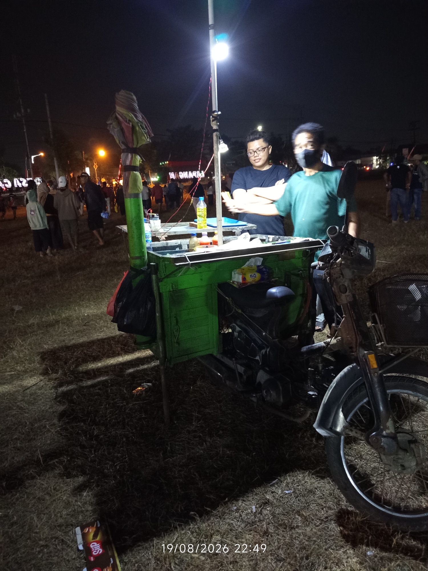 This image, captured at night in Canggu, Indonesia, depicts an outdoor scene with a food cart and several individuals. A green food cart, mounted on a motorcycle, is the central focus. The cart is illuminated by a bright overhead light. Two men are standing behind the cart; one, wearing glasses and a dark t-shirt, appears to be interacting with items on the cart. The other man, wearing a green t-shirt and a dark face mask, is facing towards the camera. Various items are visible on and around the cart, including bottles of liquid, packages, and what appears to be a rolled-up umbrella. In the background, a crowd of people are dispersed across a grassy, uneven area, suggesting a public gathering or market. The overall atmosphere is dark, with the artificial light from the cart and distant streetlights creating shadows. The timestamp in the lower right corner reads "19/08/2026 22:49".